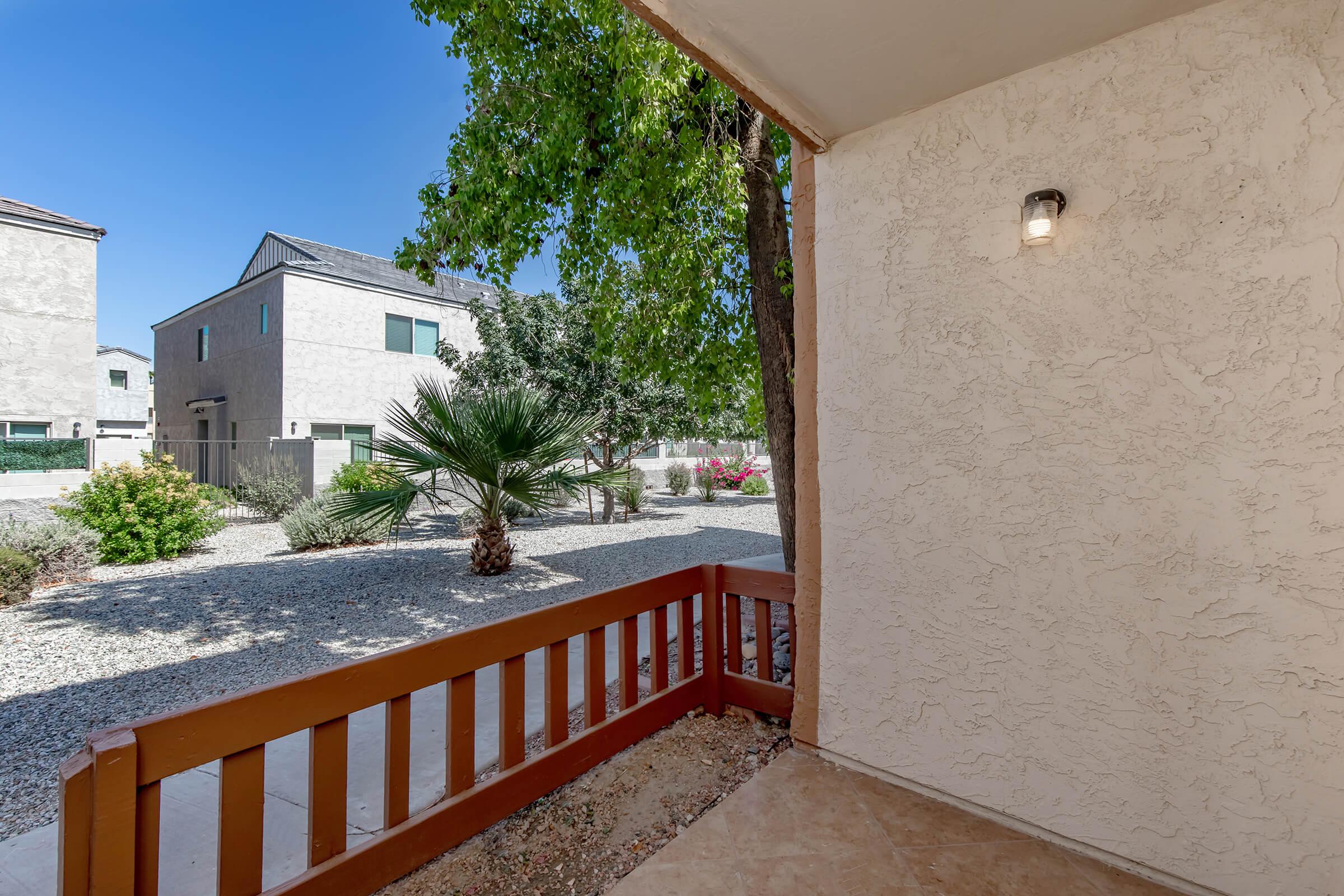 View from a covered porch showing a small outdoor space with gravel landscaping, palm trees, and several nearby buildings. Bright blue sky is visible, indicating a sunny day. The porch has a wooden railing and a light fixture on the wall.