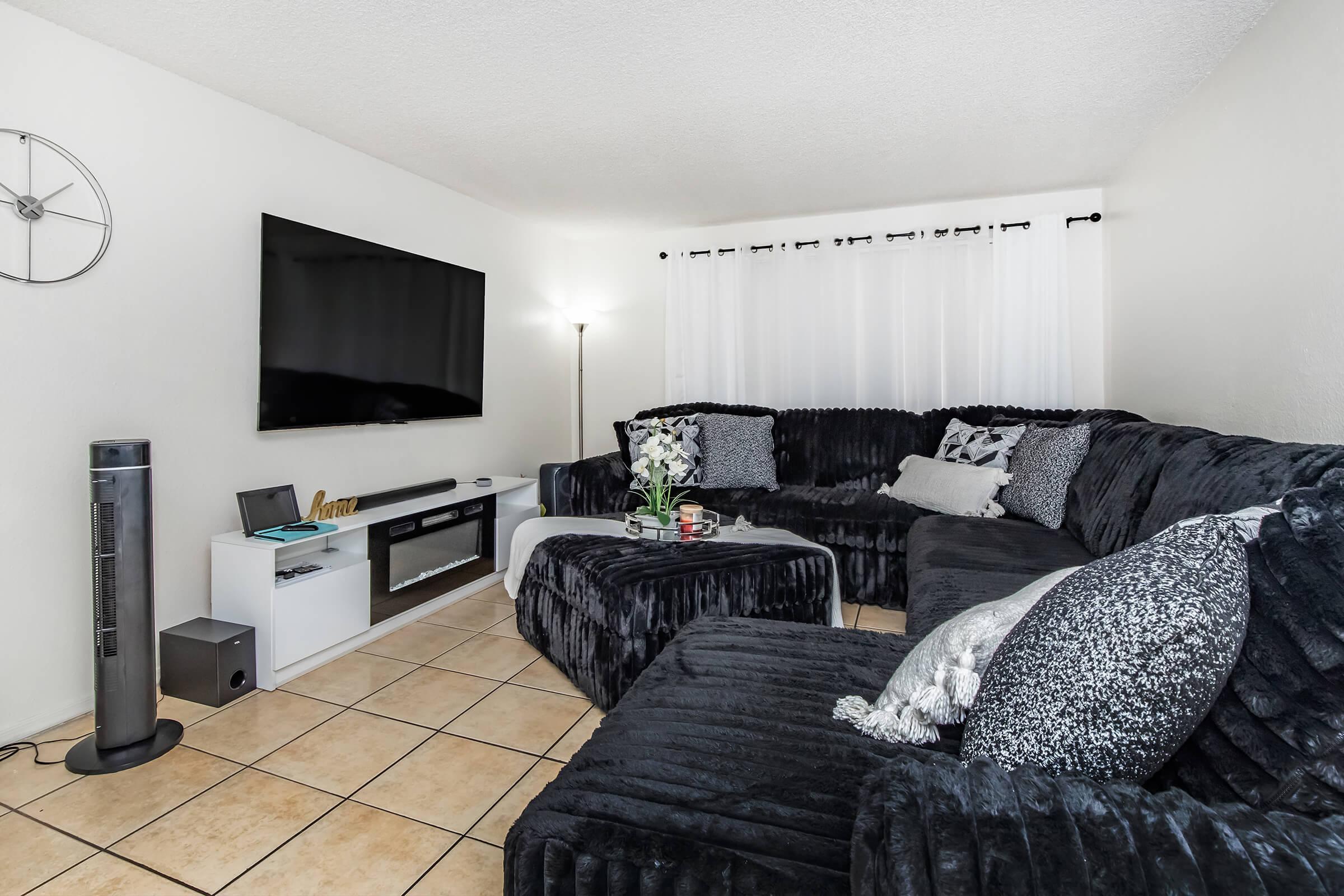 Cozy living room featuring a large dark sectional sofa with decorative pillows, a flat-screen TV mounted on the wall, a minimalist entertainment center, and a tall floor lamp. Light-colored walls and sheer curtains create a bright atmosphere, complemented by a small plant on the coffee table.