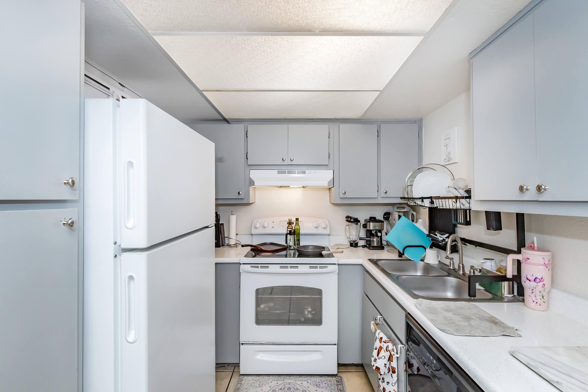 A compact kitchen featuring light gray cabinetry, white appliances including a refrigerator, oven, and microwave. There are countertops with a sink, stacked dishware, and a blue cutting board. Soft overhead lighting illuminates the space, creating a welcoming atmosphere. A small rug is visible on the floor.