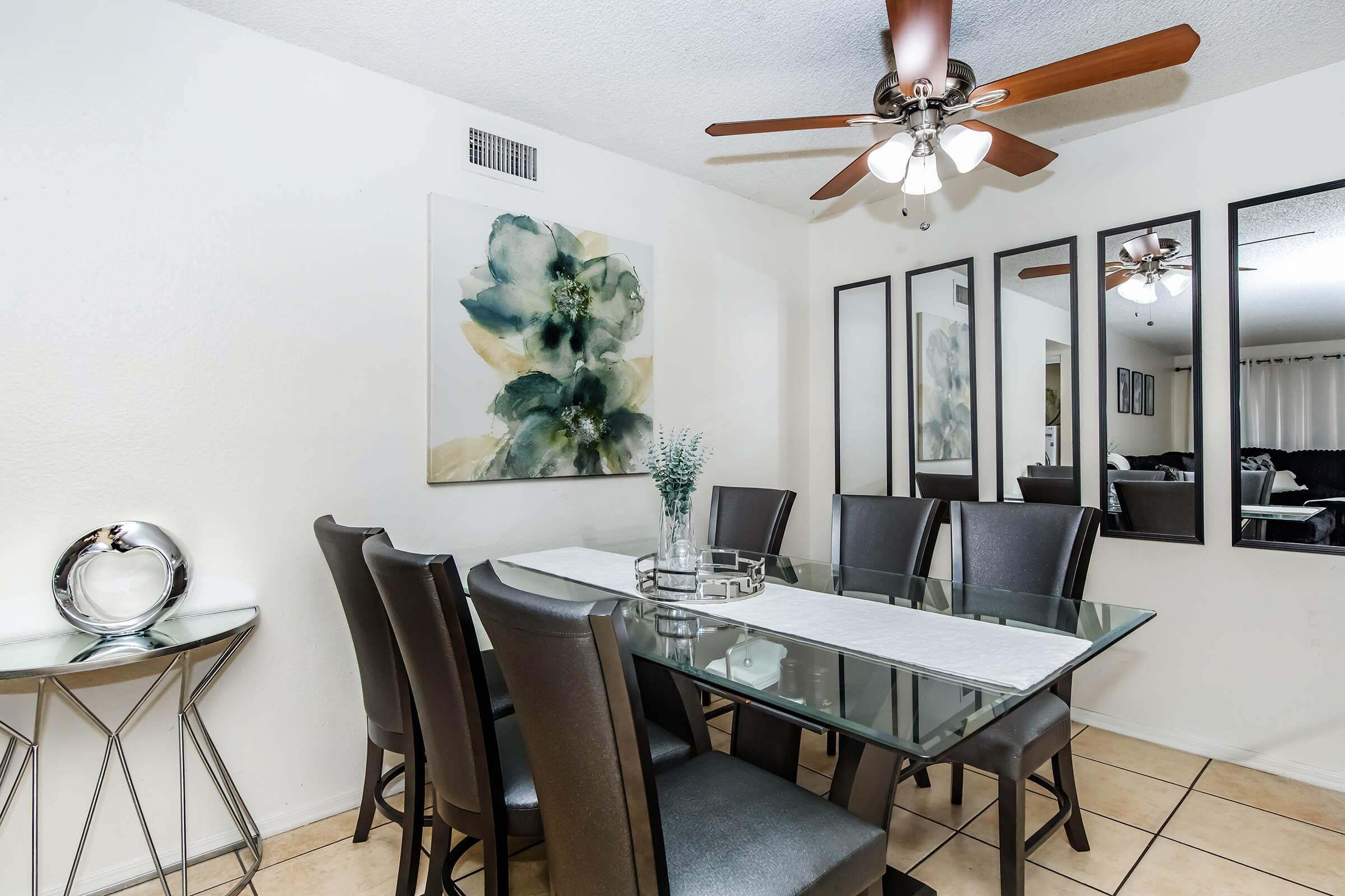 A modern dining area featuring a glass table with a white table runner, surrounded by eight black leather chairs. On the wall, there's a large floral artwork, and two mirrored panels reflect the space. A decorative silver bowl sits on a small side table. A ceiling fan provides additional lighting.