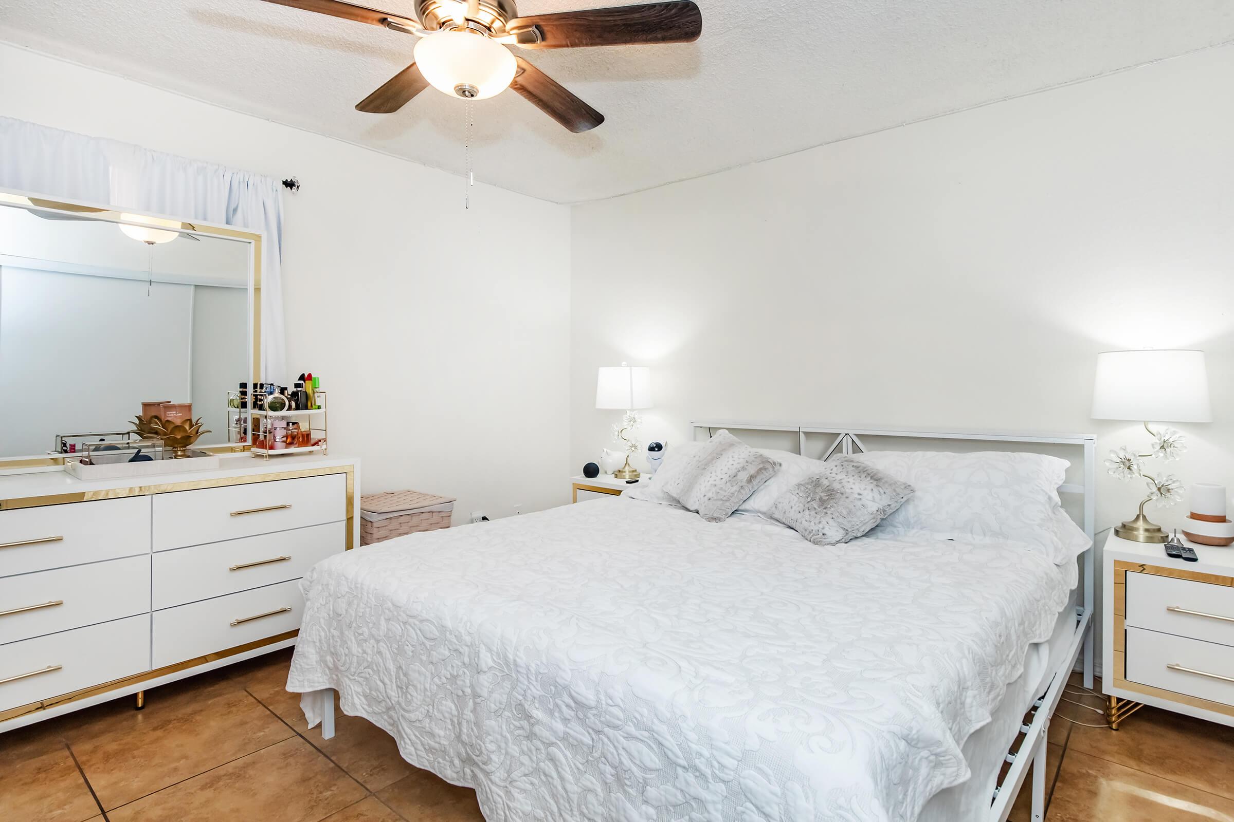 A cozy bedroom featuring a white bed with decorative pillows, a modern dresser with gold accents, two bedside lamps, and a ceiling fan. The room has neutral-colored walls and tiled flooring, with a large mirror reflecting a tidy workspace on the dresser.