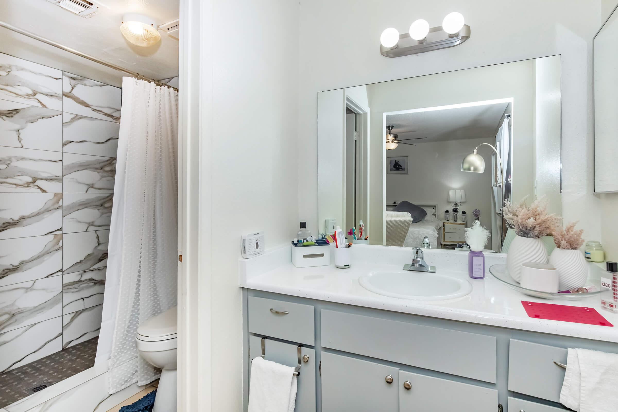 A modern bathroom featuring a white countertop with a sink, a large mirror above it, a shower area with marble-patterned tiles, and a toilet. Decor includes a small plant and toiletries on the counter. The design is bright and clean, with light-colored walls and soft lighting.