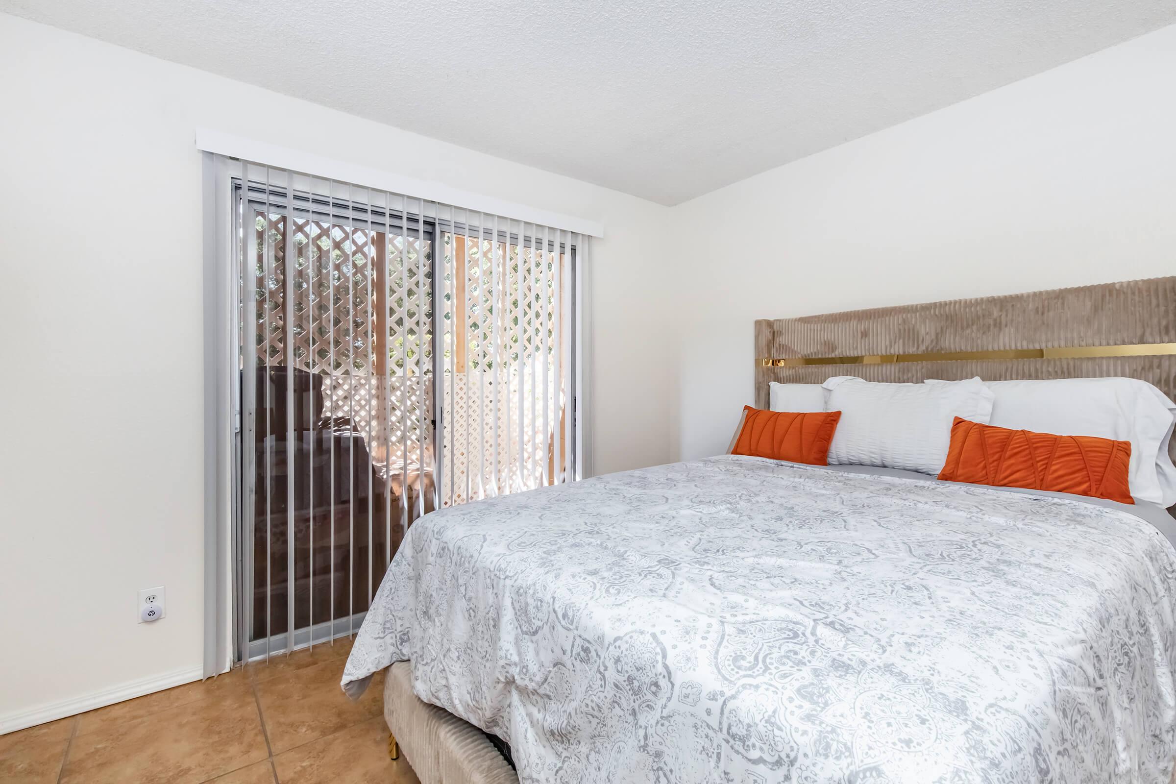A cozy bedroom featuring a neatly made bed with decorative pillows in orange and white. There is a large window with vertical blinds, allowing natural light to brighten the space. The walls are painted in a light color, and the flooring consists of beige tiles, creating a warm atmosphere.