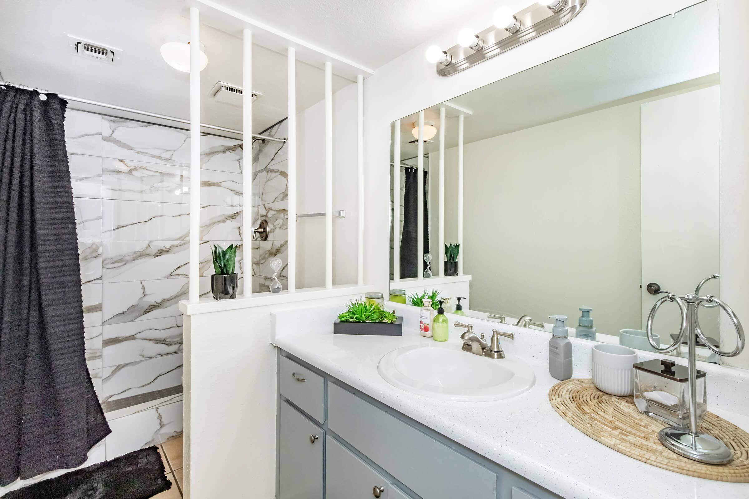 A modern bathroom featuring a white countertop with sink, a large mirror, and stylish decorations. The shower area has marble tiles and a black curtain, while green plants add a touch of nature. The overall design is clean and contemporary.
