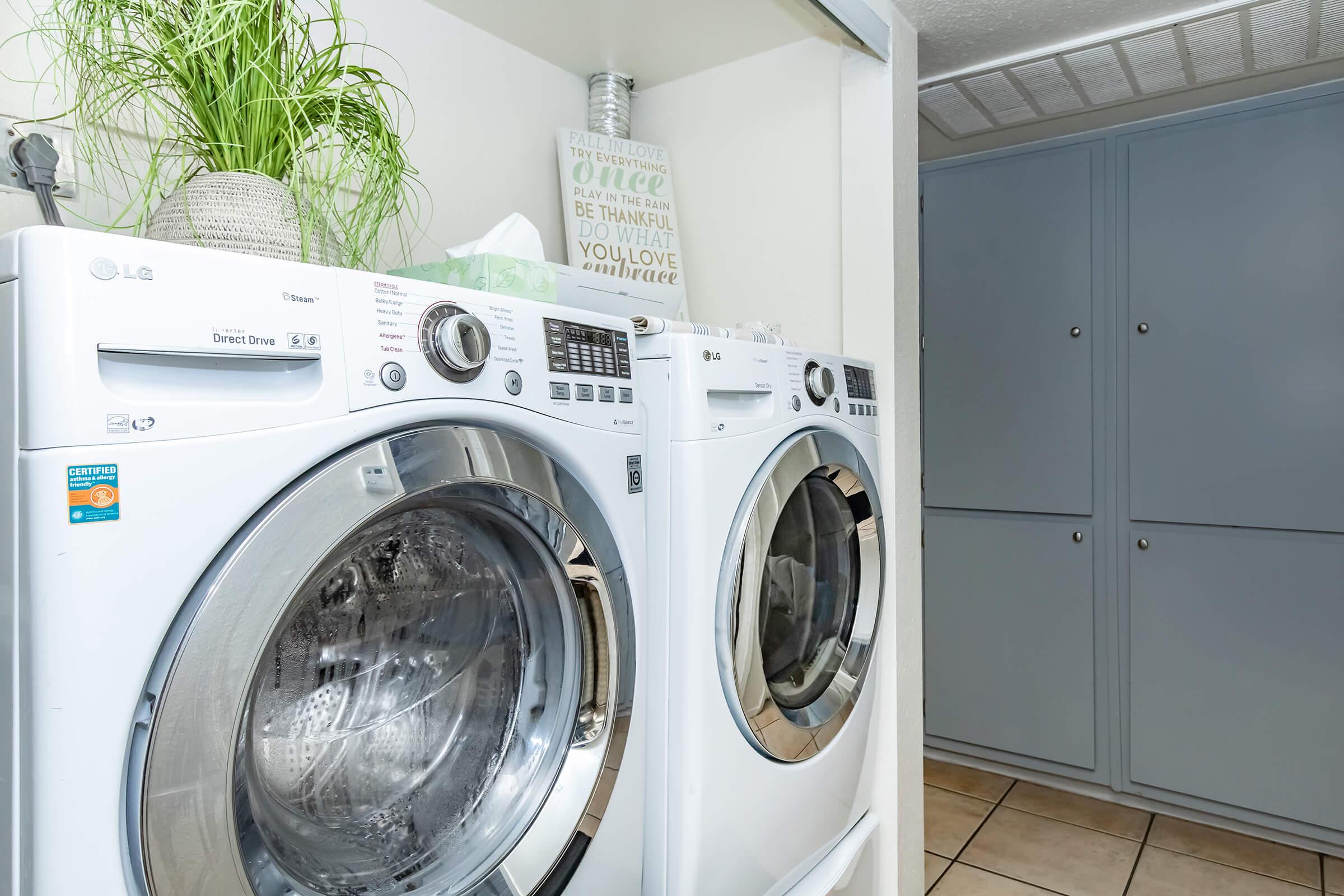 A laundry room featuring a pair of modern, white washing machines stacked side by side. Above them, a decorative plant is placed on a shelf, along with a framed sign with inspirational text. The walls are painted light colors, and there are grey cabinets in the background. The floor is tiled.
