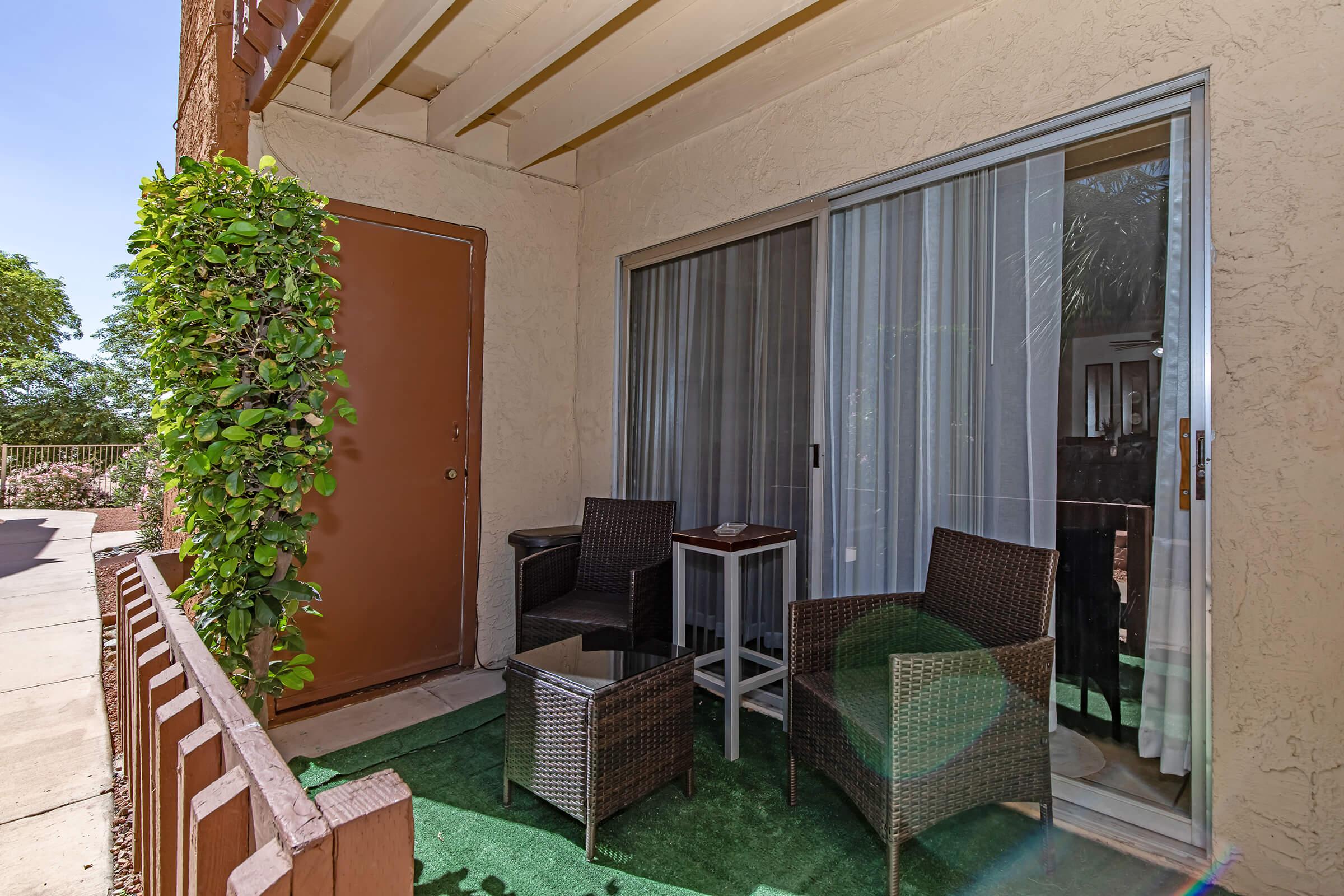 A small outdoor patio area featuring a brown door, a glass table with a decorative item, and two wicker chairs on green carpet. The patio is surrounded by leafy plants and has sheer curtains partially drawn. The setting is bright with natural light filtering in.