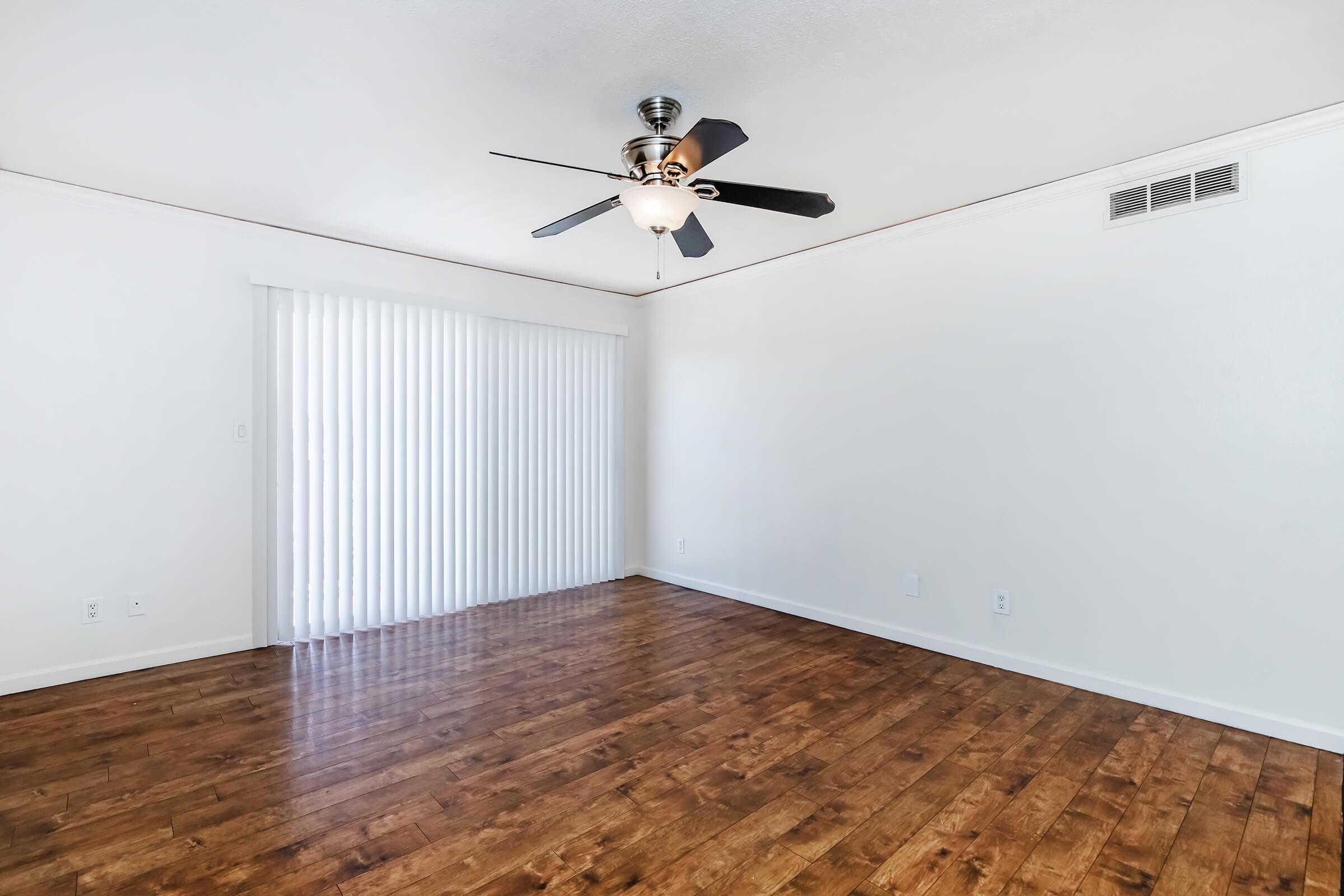 Spacious living room with a ceiling fan and large window covered by vertical blinds, allowing natural light to filter in. The floor features polished wood, enhancing the bright and airy atmosphere of the room. The walls are painted a light color, creating a neutral backdrop for decor.