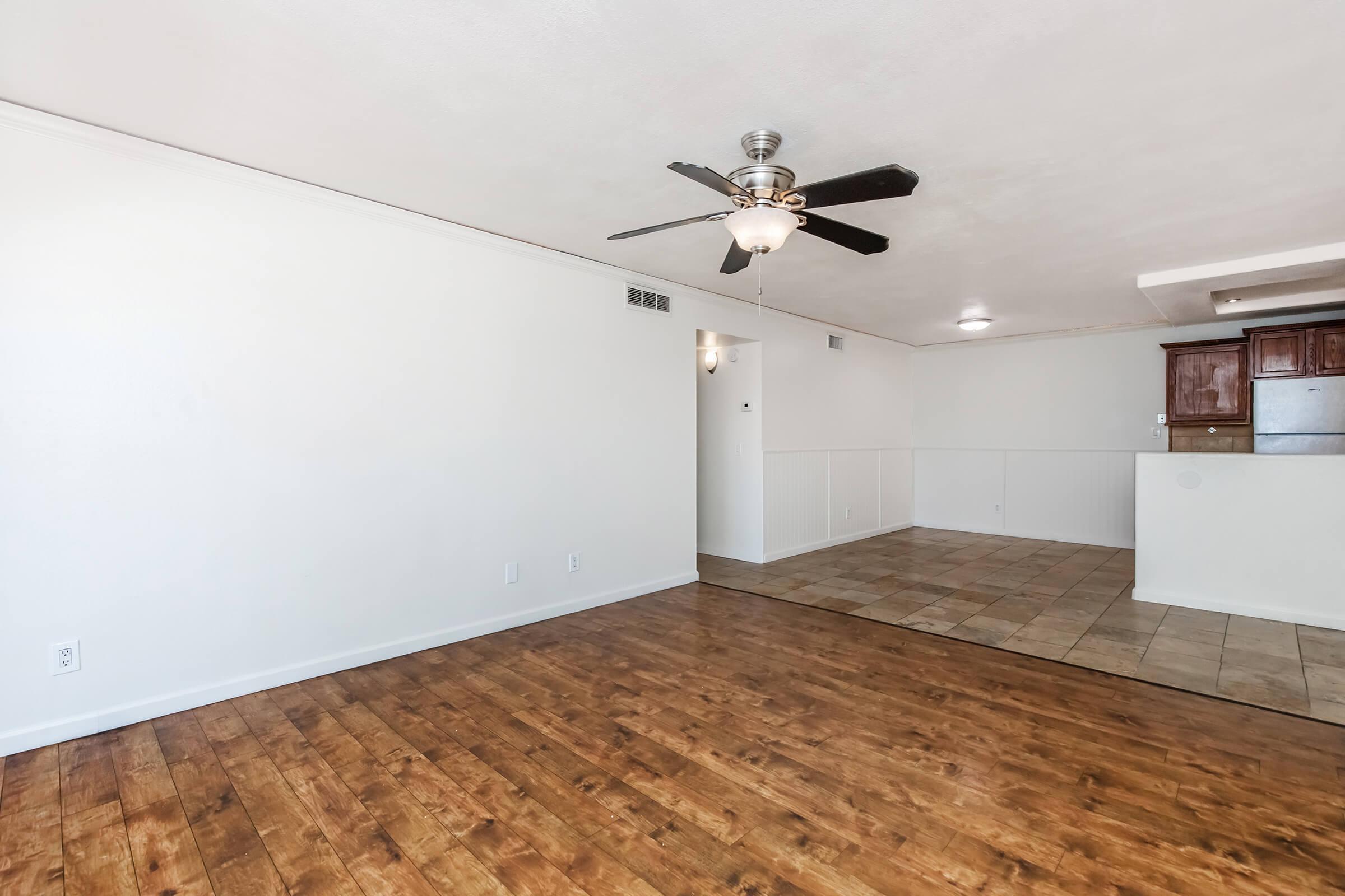 An empty living room with hardwood flooring and a ceiling fan. There is a wall with a textured finish and a doorway leading to another room. To the right, a tiled area transitions into a kitchen space with wooden cabinets visible in the background. The room is well-lit with natural light.
