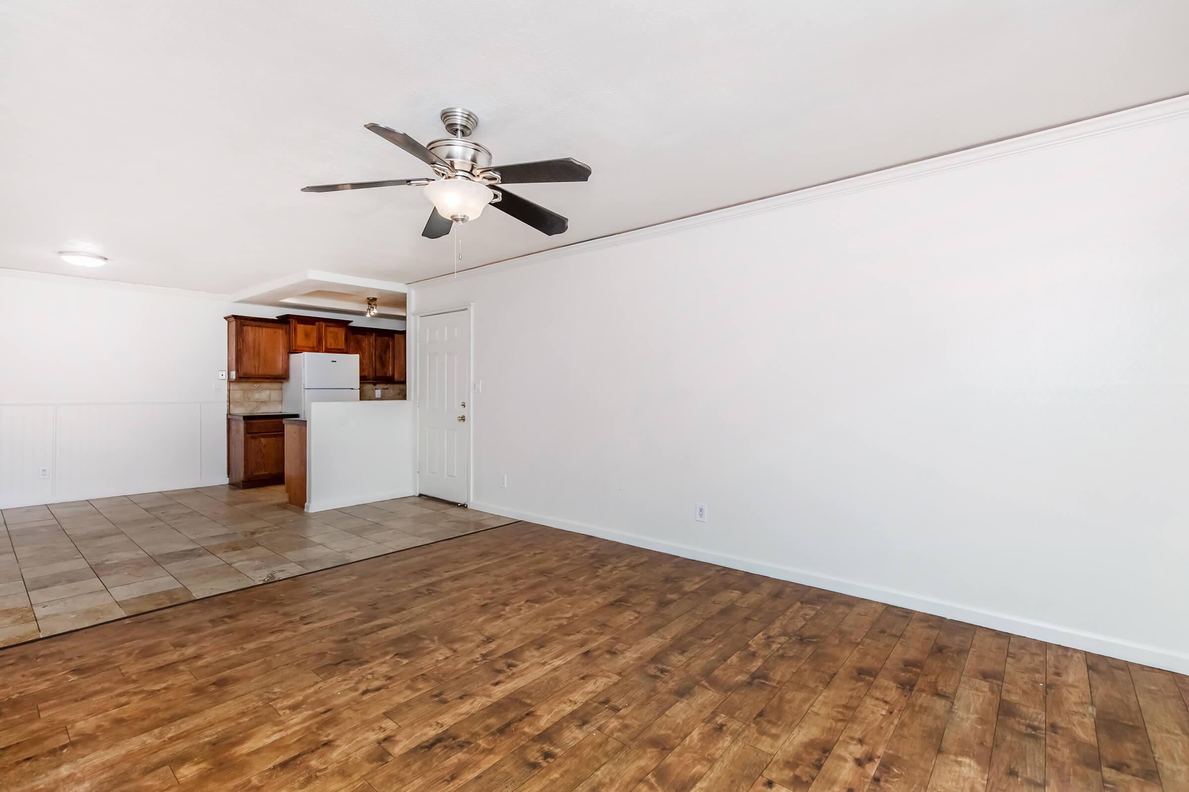 Interior of a living space featuring a ceiling fan, hardwood-style flooring, and a light-colored wall. In the background, there is a kitchen area with wooden cabinets and a white refrigerator. The room is well-lit and appears spacious.