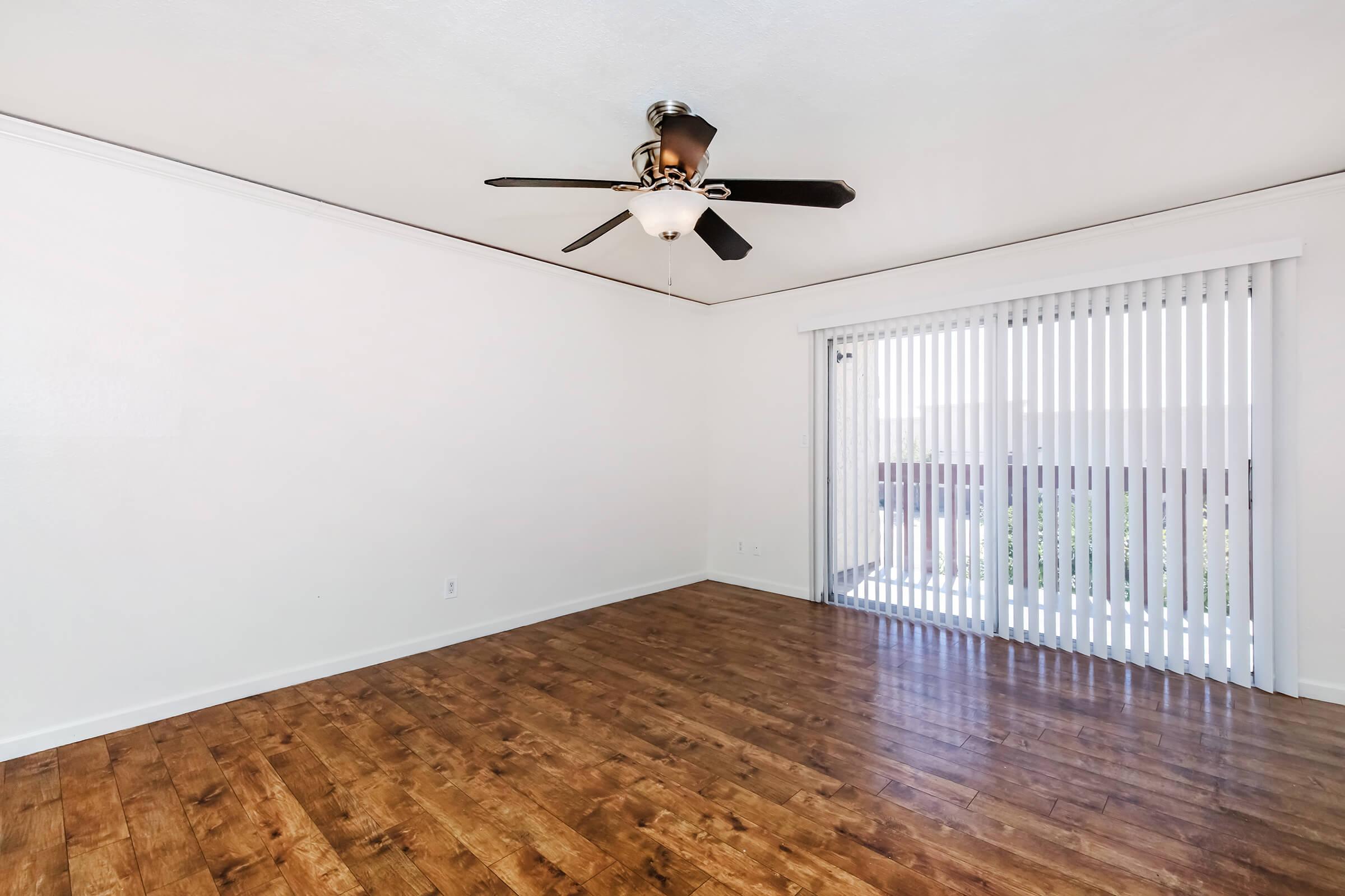 A spacious, airy room with light-colored walls and a wooden floor. A ceiling fan is mounted in the center, and there's a large sliding door with vertical blinds that leads to a balcony or patio area, allowing natural light to illuminate the space.
