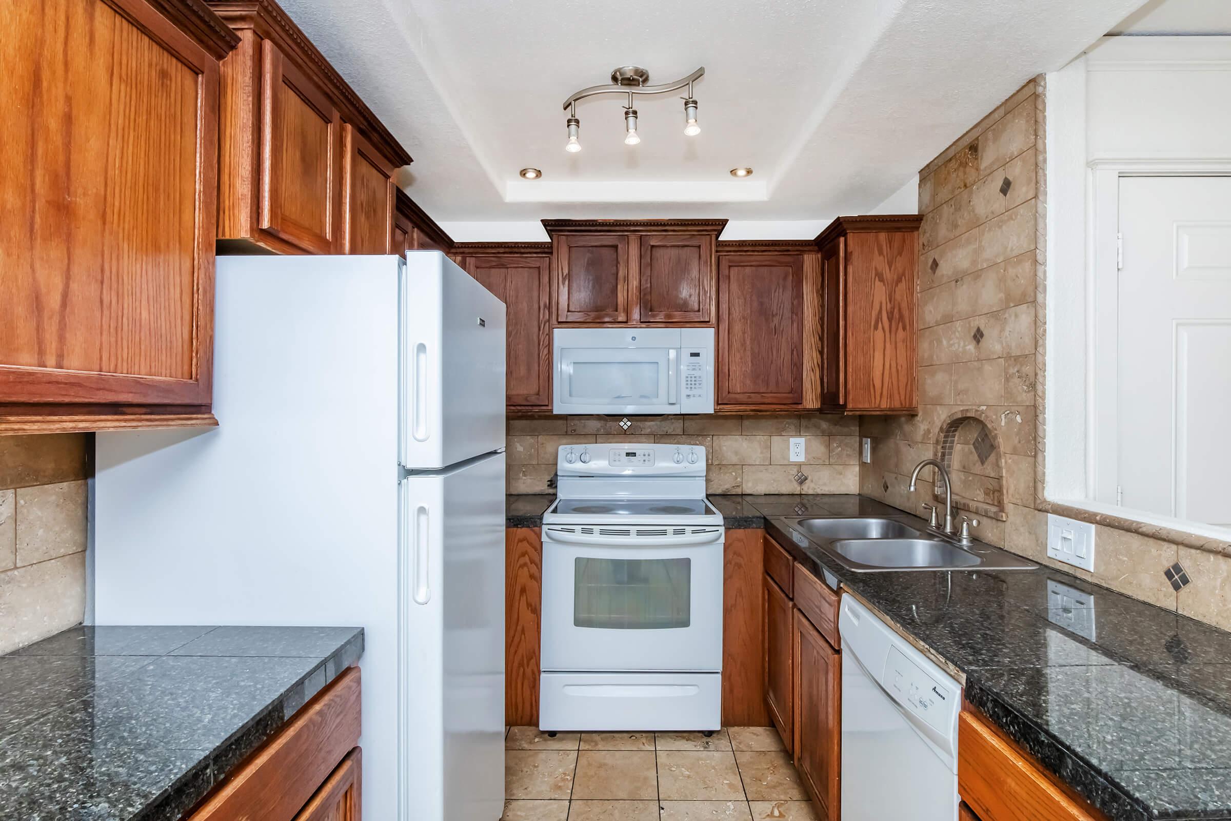 A stylish kitchen featuring dark wood cabinets, granite countertops, and modern appliances including a refrigerator, oven, microwave, and dishwasher. The space is well-lit with recessed lighting and has beige tiled walls, creating a warm and inviting atmosphere.