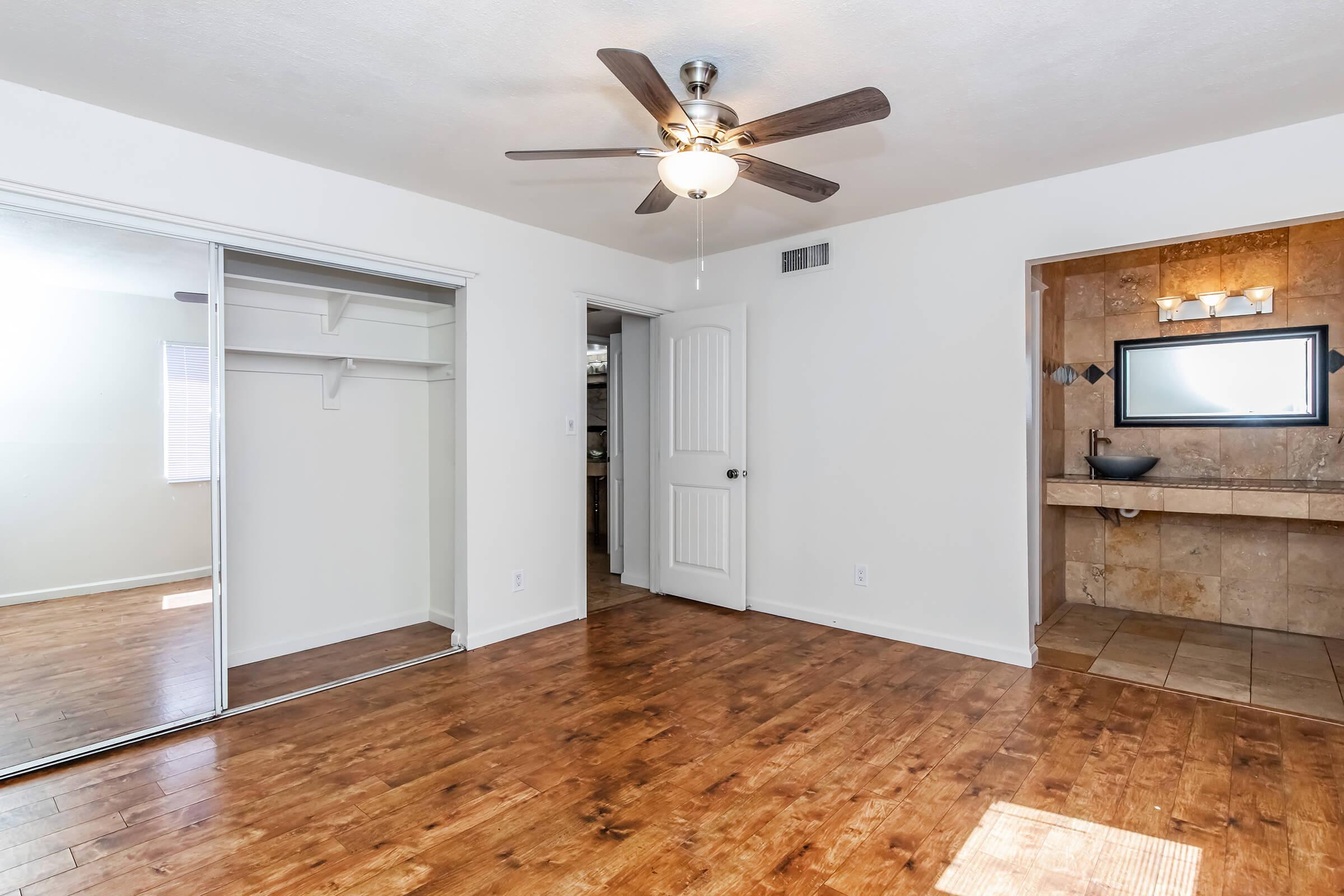 Spacious bedroom featuring a ceiling fan, hardwood flooring, and a large mirror closet. The room includes a door leading to another area and a wall with a stone accent where a modern sink and mirror are displayed. Natural light enters through the window, creating a bright and inviting atmosphere.