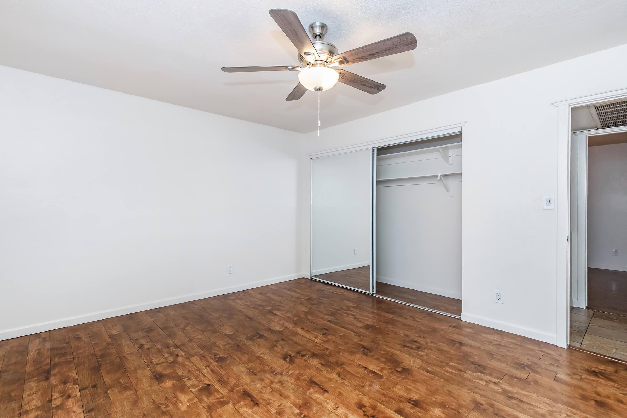 A spacious, empty room with light-colored walls and hardwood flooring. It features a ceiling fan with light, a mirrored closet with sliding doors, and an adjacent doorway leading to another space. The room is well-lit and has a clean, modern appearance.