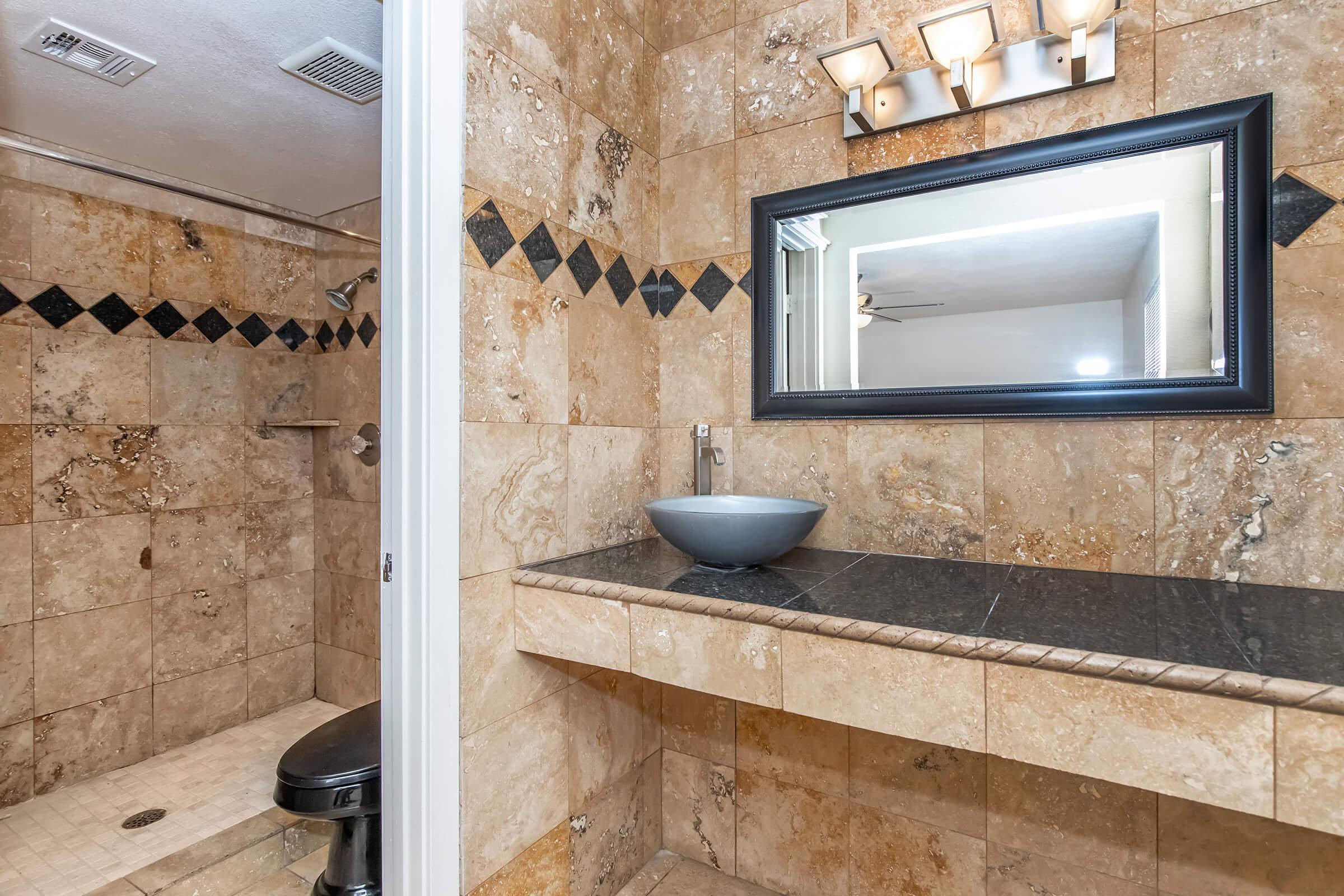 A modern bathroom featuring beige stone walls, a large mirror with a black frame, and a vessel sink on a dark countertop. The shower area is visible with a tiled floor and wall, and a sleek light fixture above the mirror. The design combines elegance with functionality.