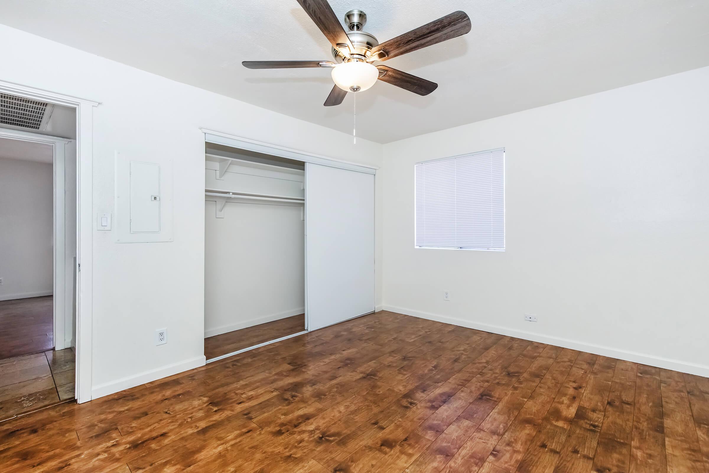 A spacious, empty room with hardwood flooring, featuring a ceiling fan and a window with blinds. There's a sliding closet door on one wall, and a doorway leading to another area is visible. The walls are painted white, creating a bright and airy atmosphere.