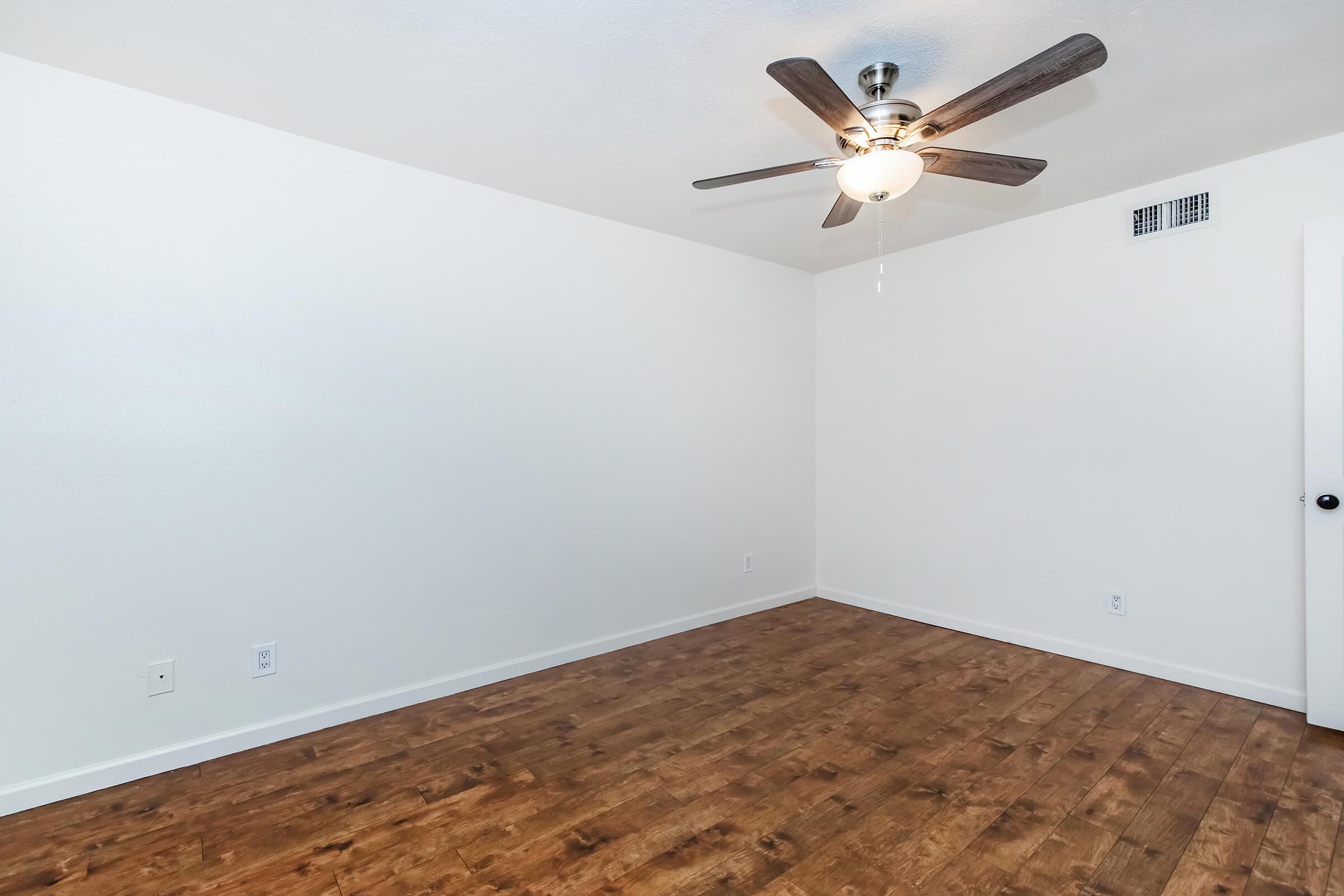 A well-lit empty room featuring a ceiling fan, light fixture, and wooden flooring. The walls are painted white, and there is a door on the right side. The room has a spacious feel with no furniture, ideal for personal customization.