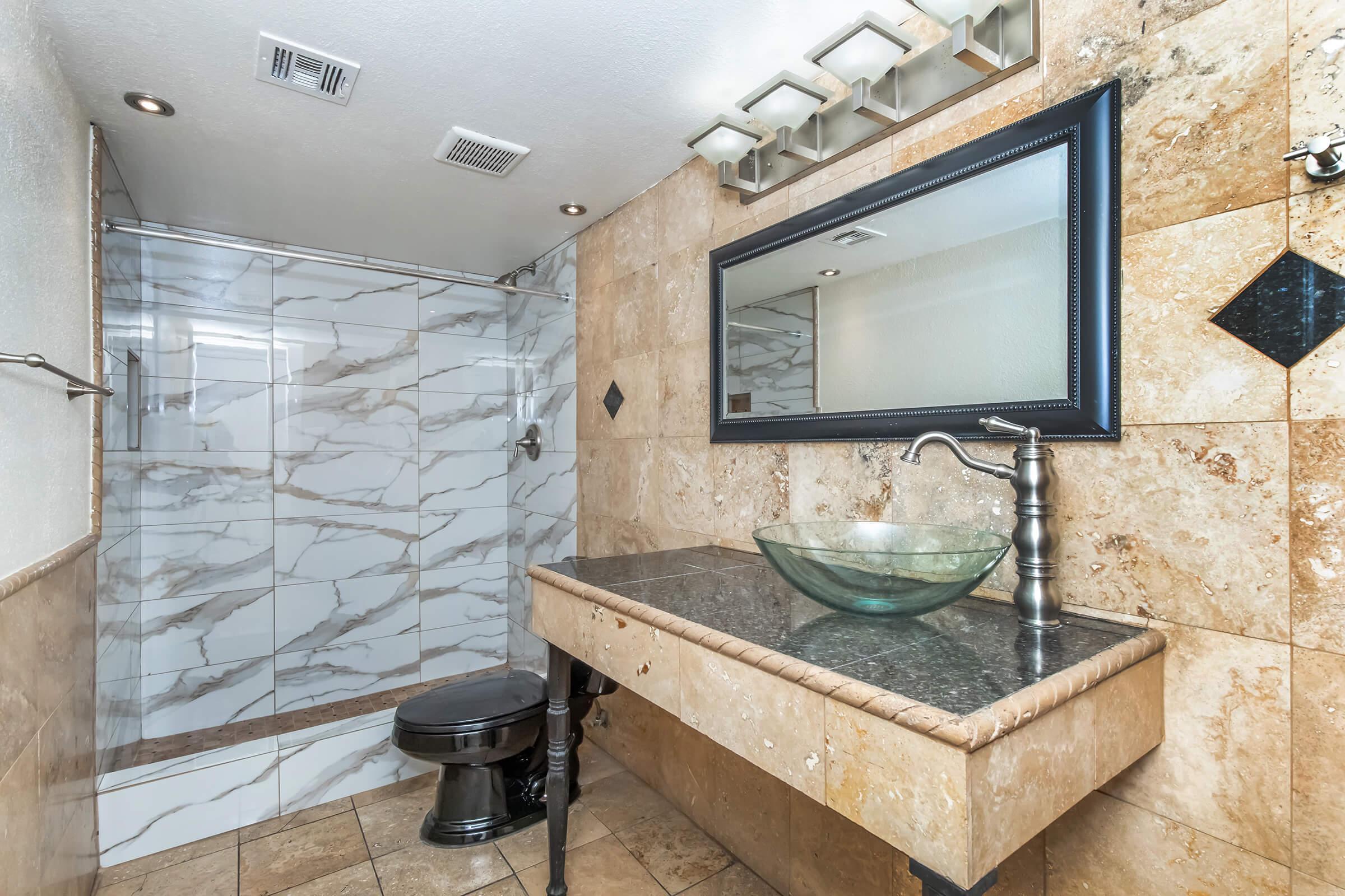 A modern bathroom featuring a spacious shower with marble tiles, a sleek black toilet, and a stylish vanity with a glass sink. The walls are adorned with light-colored stone, and there's a large mirror above the vanity, complemented by unique light fixtures.