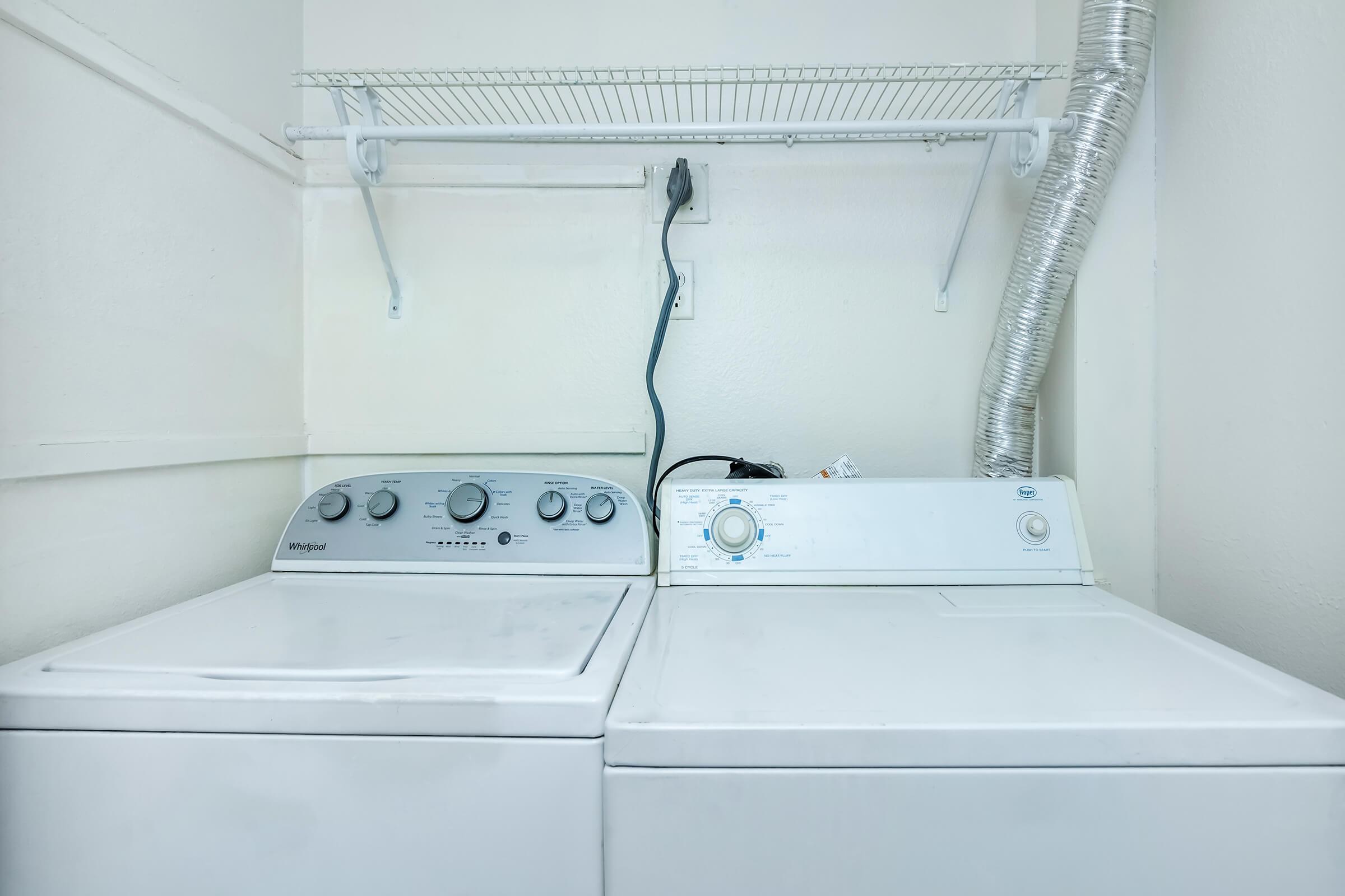 A laundry room featuring two washing machines side by side: a top-loading model on the left and a front-loading model on the right. Above them, there's a wire shelf for storage, and a vent pipe is visible on the wall. The space is well-lit with a clean, minimalist design.