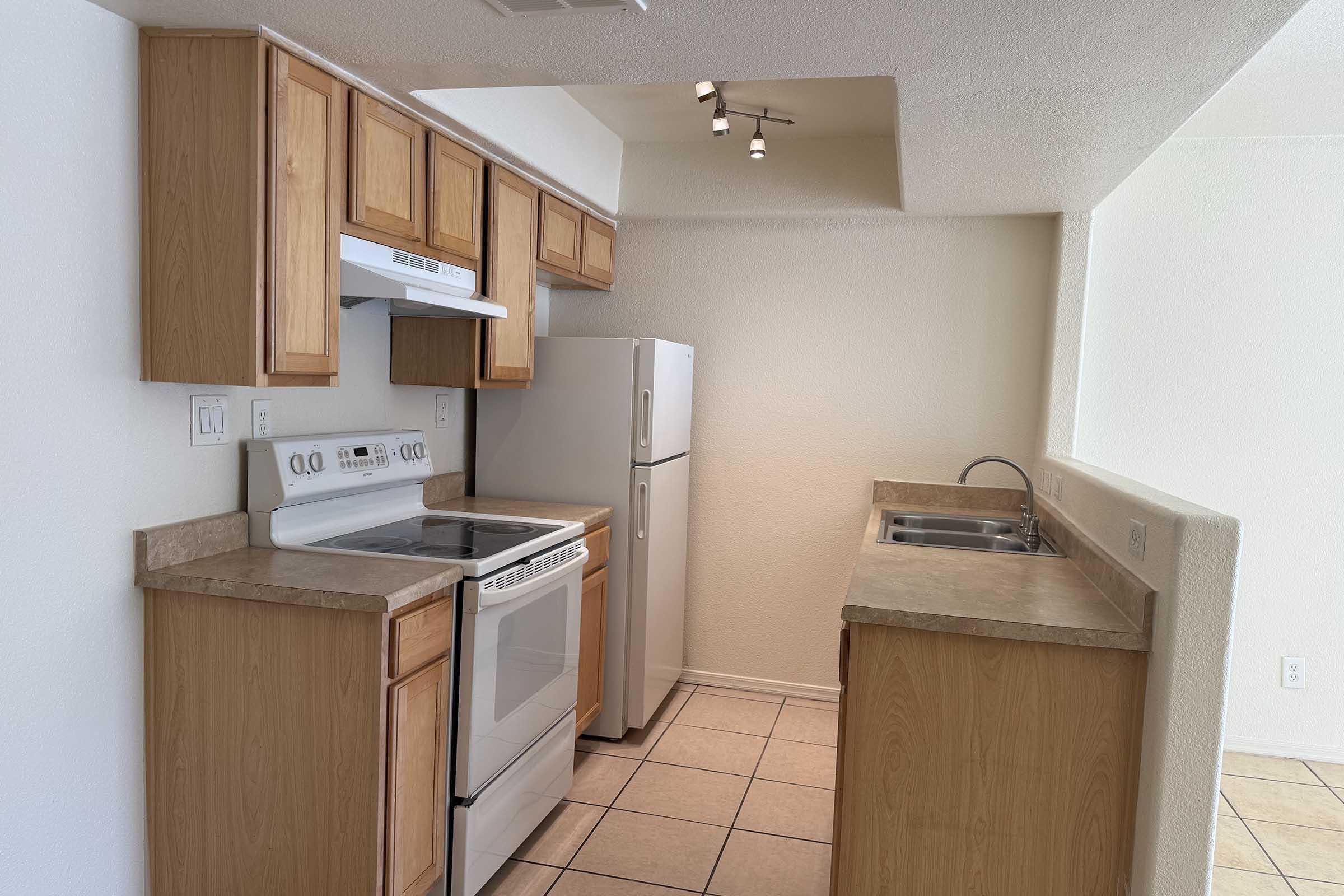 Updated kitchen featuring light wood cabinetry, a white stove and oven, and a stainless steel refrigerator. The countertop is light-colored with a single-basin sink and a small window, providing natural light. The floor is tiled, and the walls are a neutral shade.