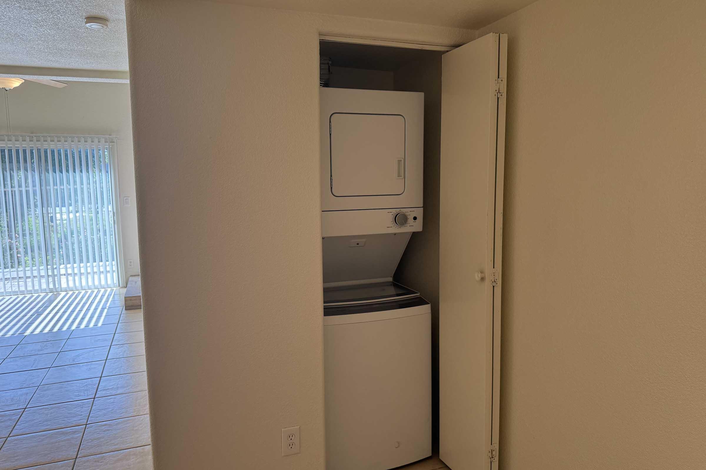 A compact laundry closet featuring a stacked washer and dryer unit, with a partially open door. The background shows a glimpse of a living area with tiled flooring and a sliding glass door leading to an outdoor space.
