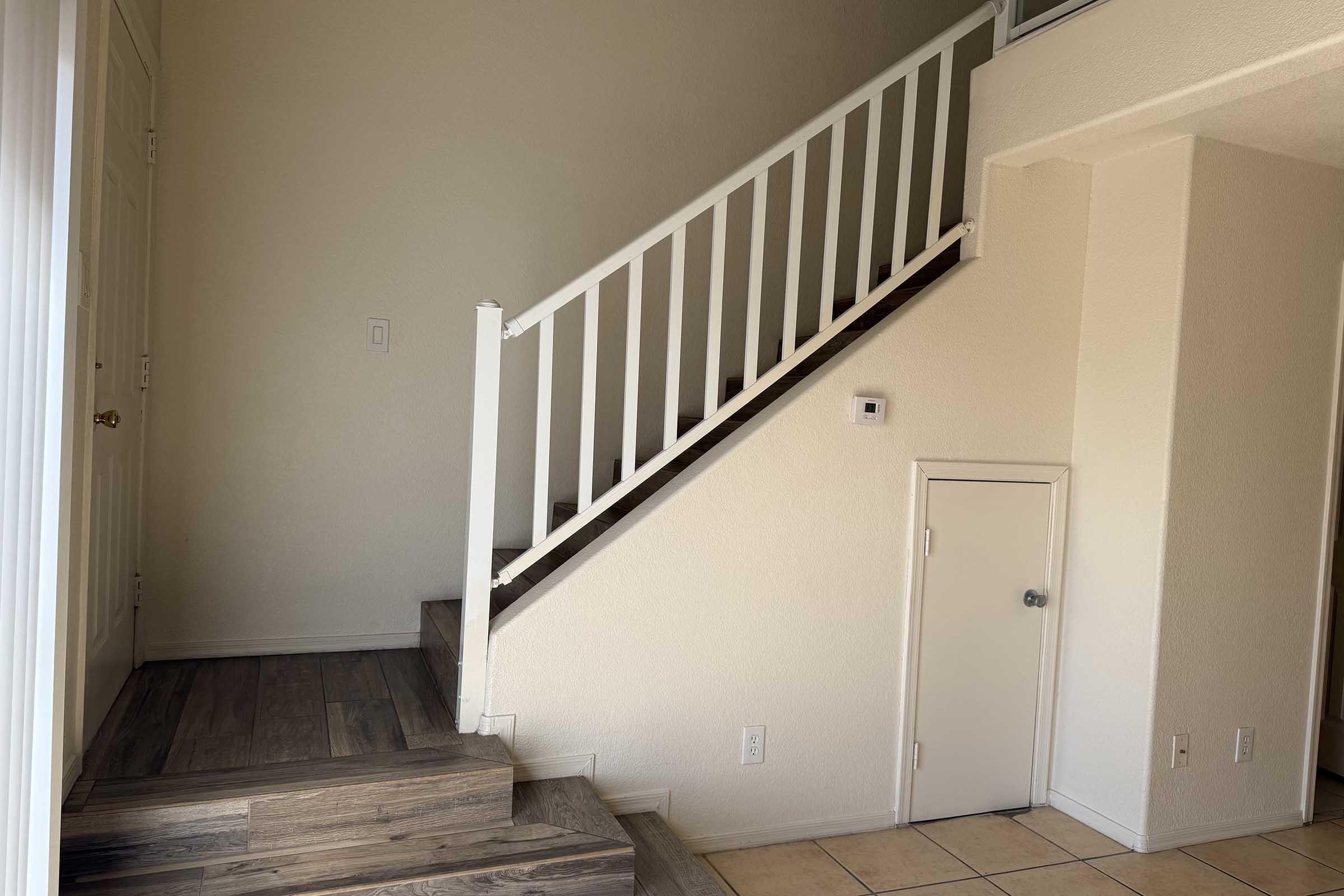 A well-lit foyer featuring a staircase with a white railing, leading to an upper level. The walls are painted a soft beige, and there is a small door beneath the stairs. The flooring consists of light-colored tiles near the entrance and wooden steps leading up.