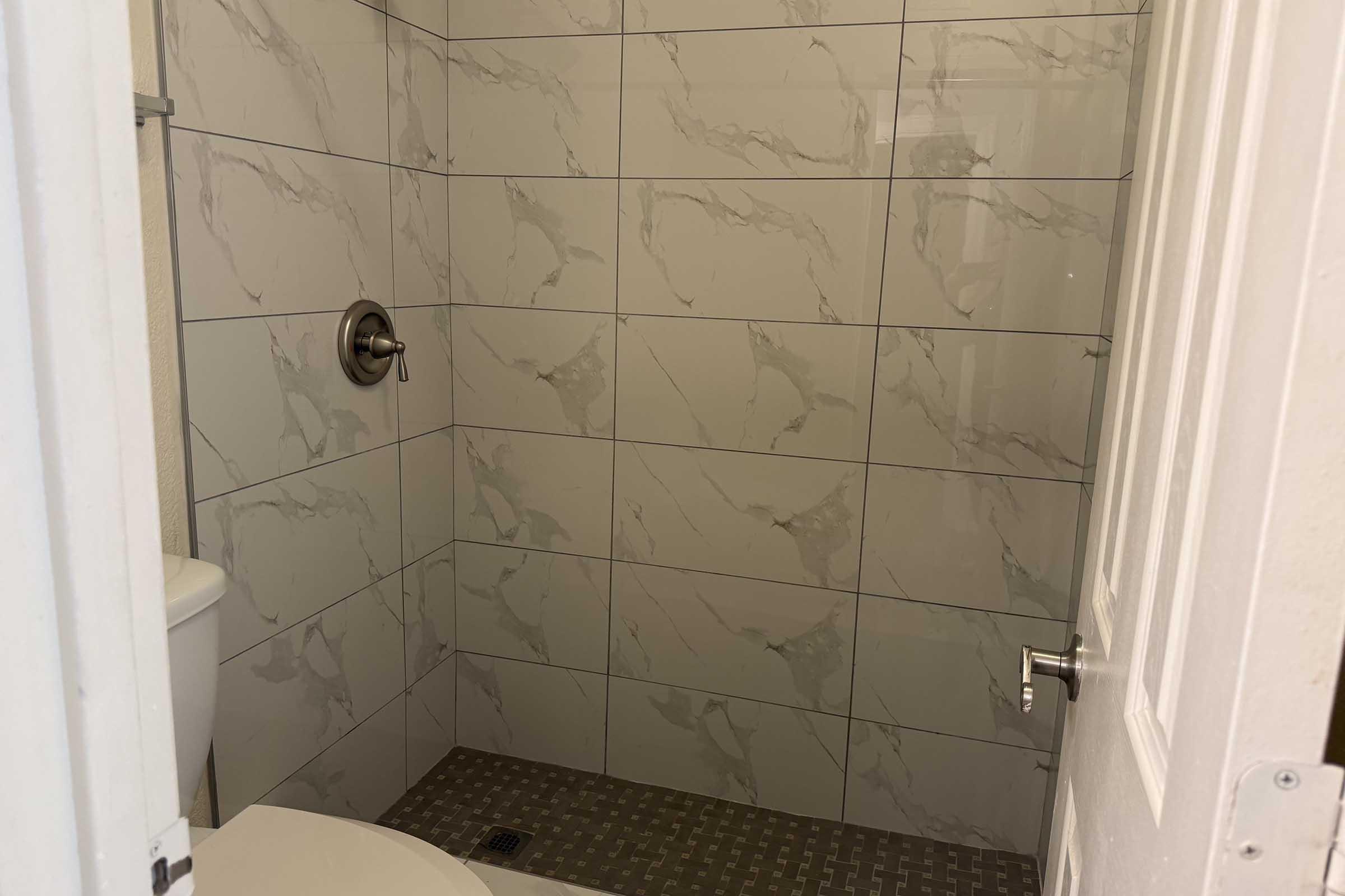 A bathroom shower area featuring marble-patterned tiles on the walls, a silver shower handle, and a tiled floor. A white toilet is partially visible to the left, and a white door is closed on the right side of the image.