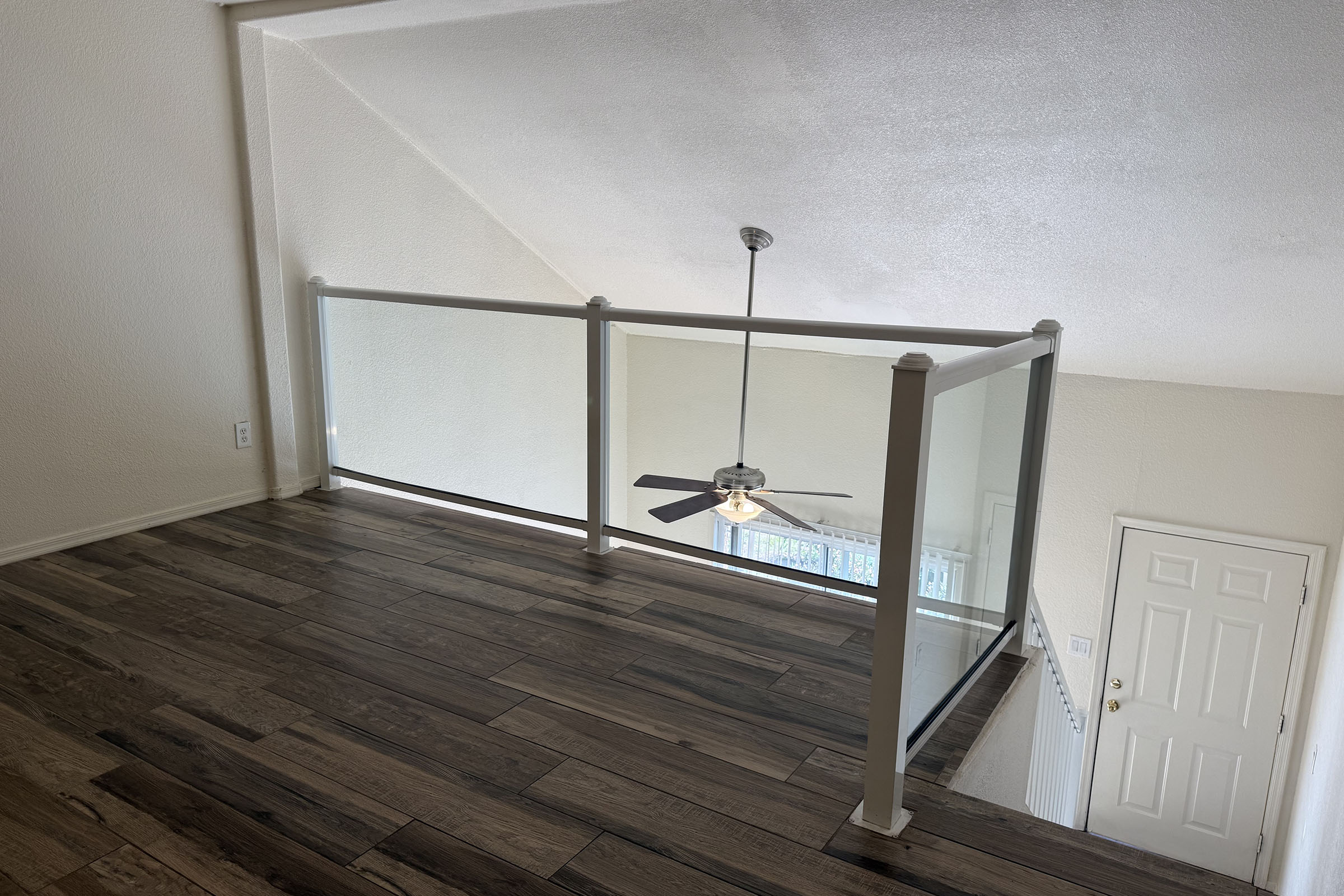 Interior view of a modern loft space featuring high ceilings, a glass railing, a ceiling fan, and wooden flooring. The entrance door is visible in the background, along with natural light coming through the upper windows. The overall decor is simple and contemporary.