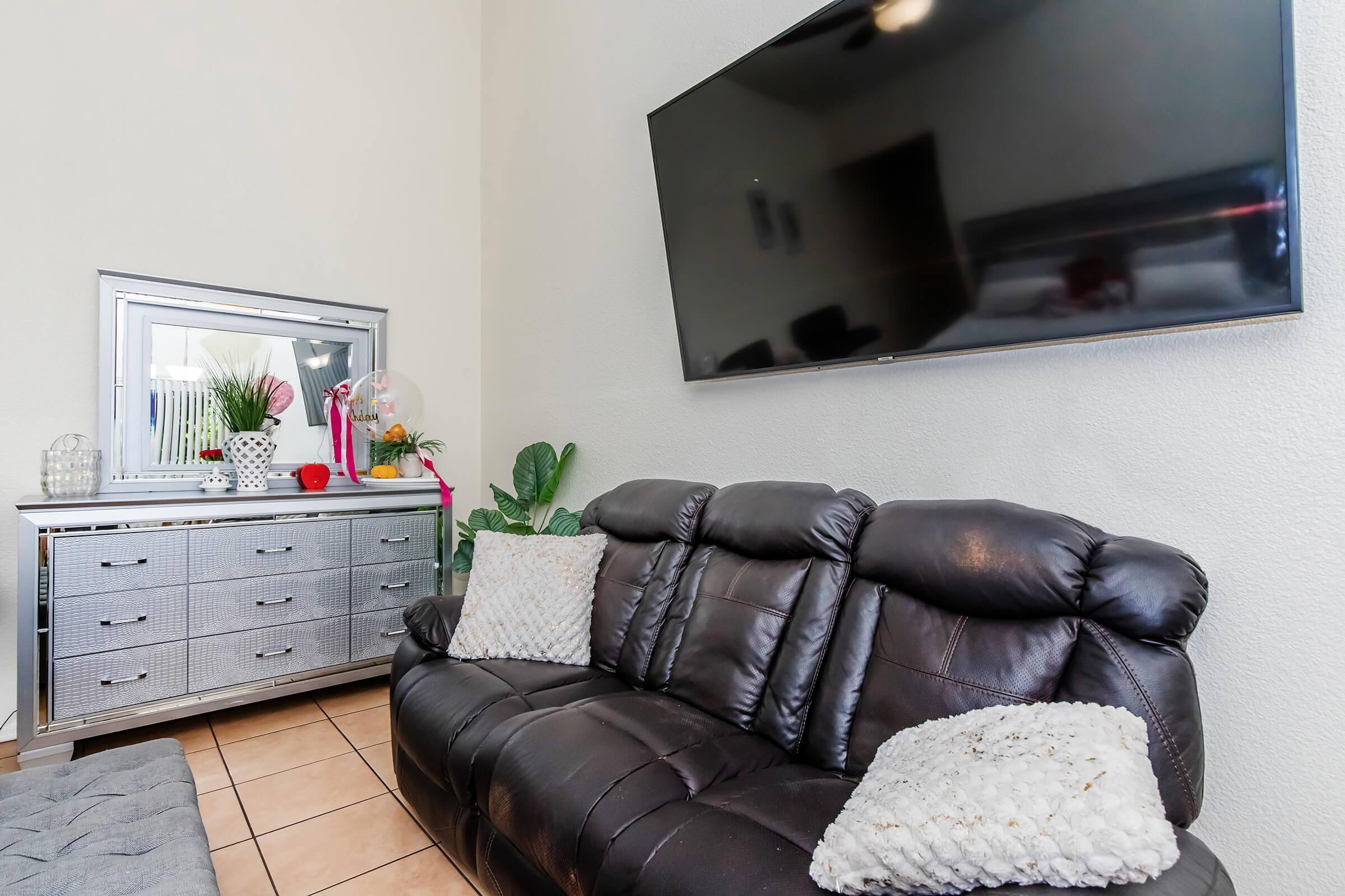 A modern living room featuring a black leather couch with decorative pillows, a silver mirrored dresser, and a wall-mounted flat-screen TV. There are plants and decorative items on the dresser, adding a touch of color to the neutral walls and tiled floor.
