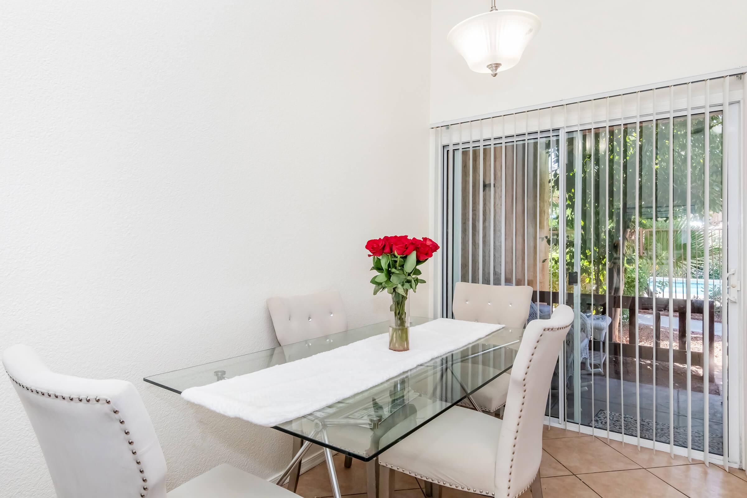 A modern dining area featuring a glass table with a white runner and a vase of red roses. Surrounding the table are four light-colored chairs. An open sliding door offers a view of greenery outside, with natural light illuminating the space. The walls are painted white, creating a bright atmosphere.