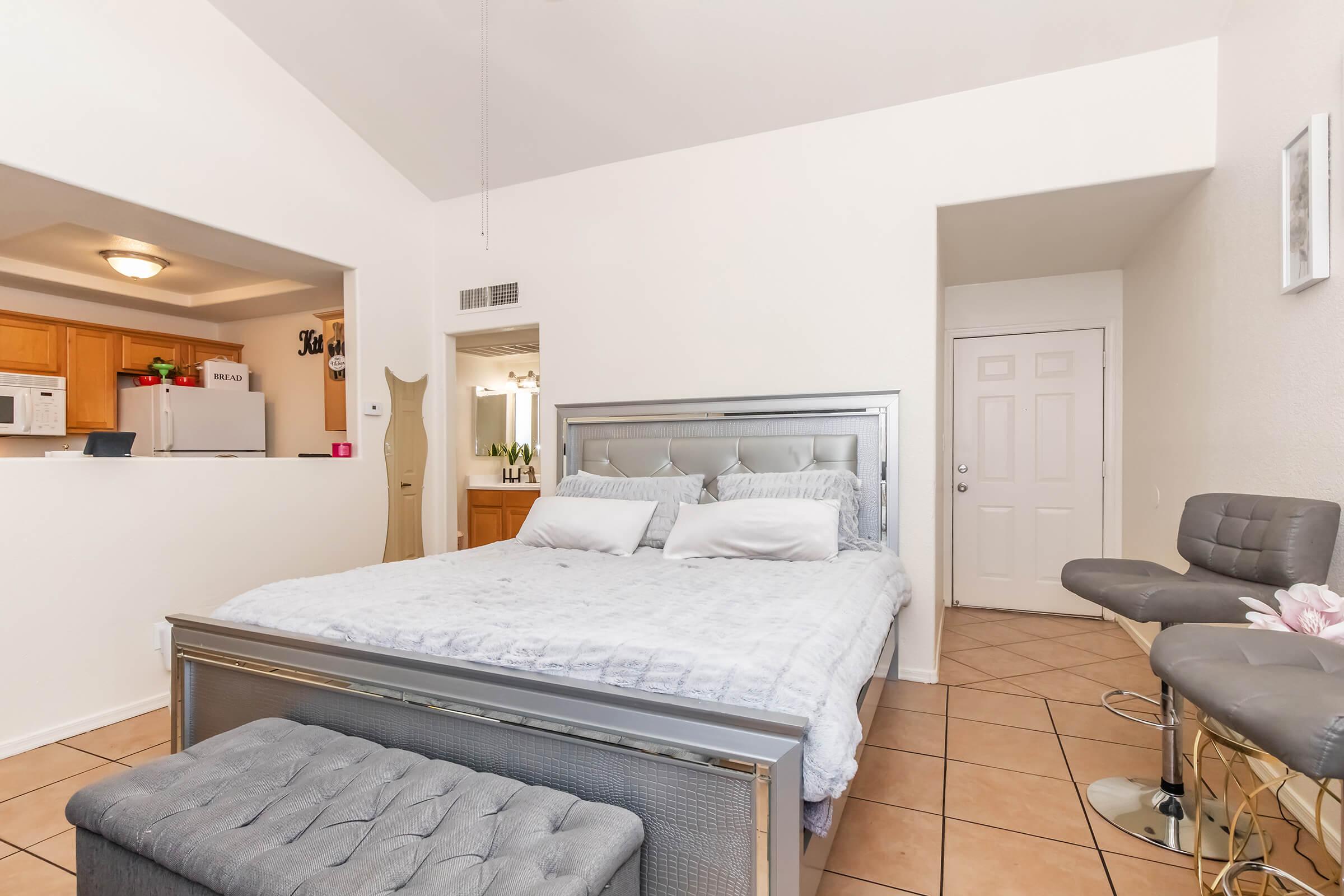 A cozy bedroom featuring a large bed with gray bedding, two pillows, and a tufted bench at the foot. The room has a light color scheme, tiled flooring, and a high ceiling. In the background, a small kitchen area with bar stools is visible, creating a modern and inviting atmosphere.