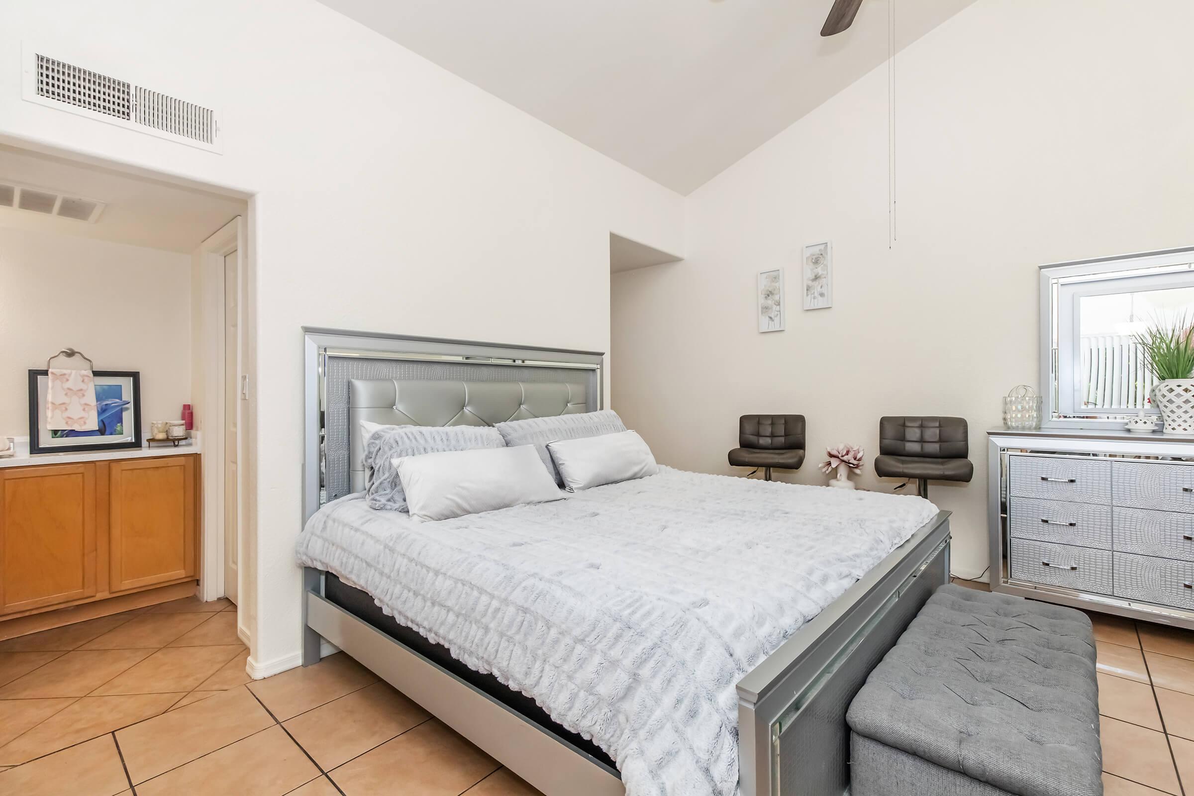 A modern bedroom featuring a silver bed frame with a plush gray blanket and pillows. There are two stylish chairs beside the bed and a mirrored dresser. The room has light-colored walls and tiled flooring, with decorative wall art and a ceiling fan adding to the contemporary aesthetic.