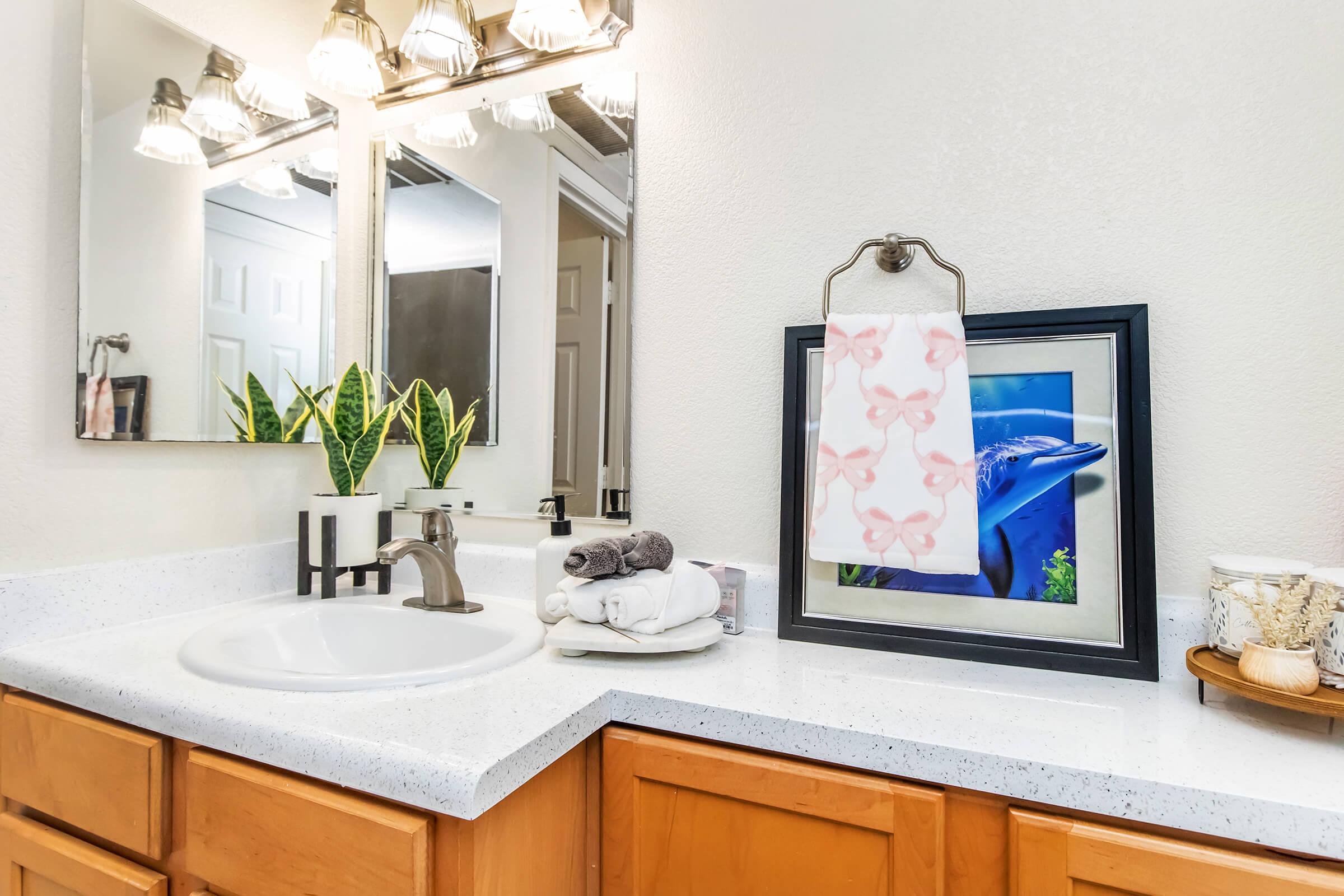 A well-lit bathroom with a sink and wooden cabinets. The countertop features neatly stacked white towels, a decorative framed photo of a blue fish, and a towel hanging on a wall-mounted holder. There are also green plants and a small decorative container on the surface, adding a touch of nature to the space.