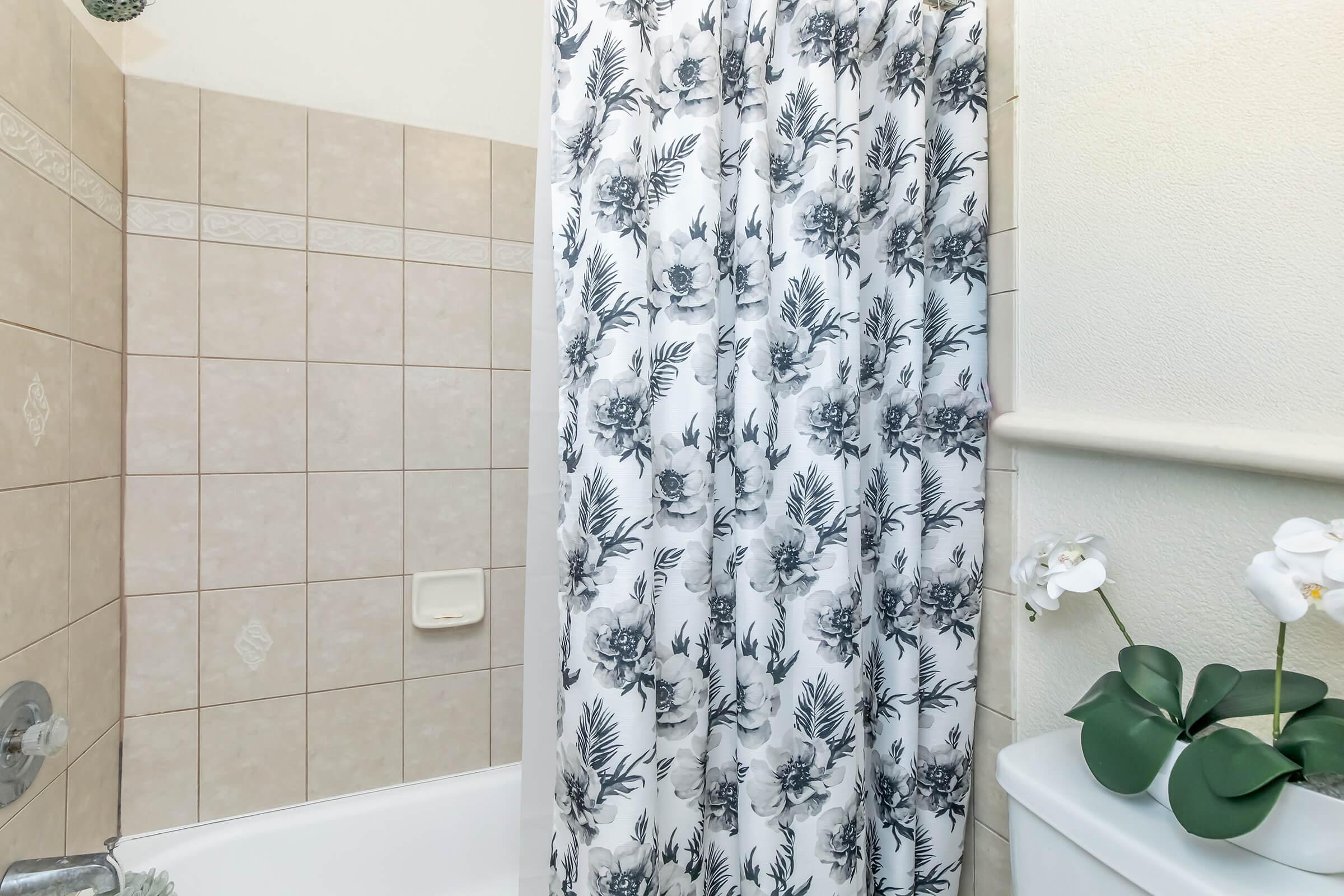 A bathroom with a bathtub and a floral shower curtain. The tiles are light beige, and there is a white toilet beside the bathtub, adorned with a small plant. The overall ambiance is clean and modern.