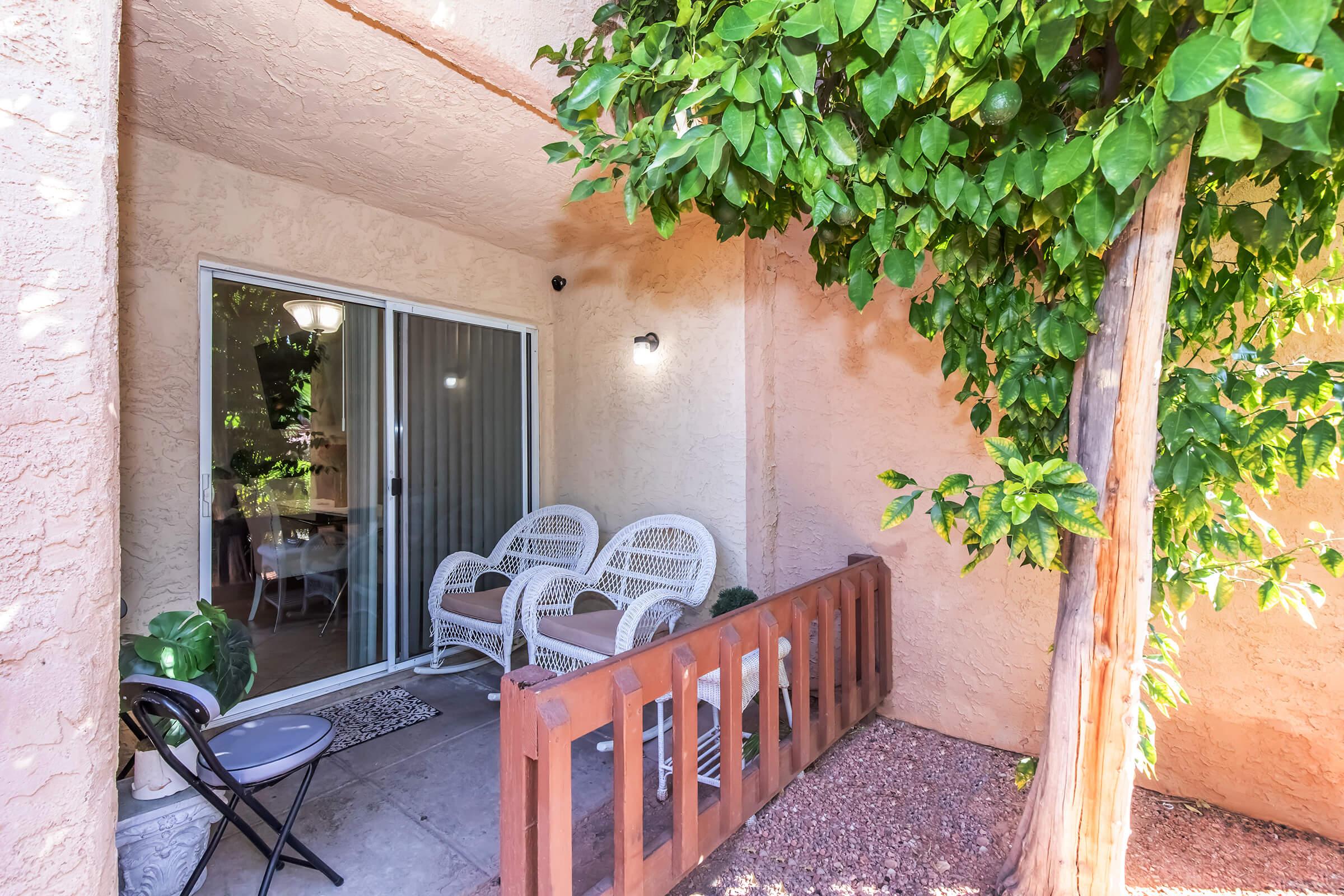A cozy outdoor patio featuring two white wicker chairs and a small round table. The area is framed by a wooden railing and lush green plants, with a large tree nearby. A sliding glass door leads into a bright indoor space, suggesting a warm and inviting atmosphere.
