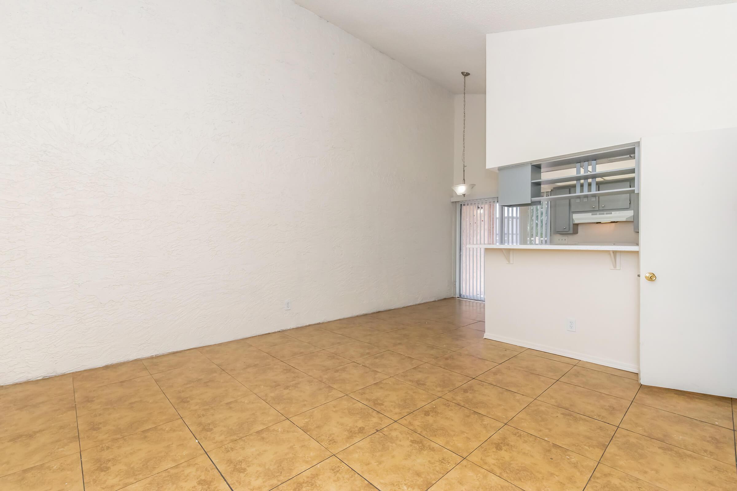 A spacious, vacant room with high ceilings and textured white walls. The floor is covered with large beige tiles. On one side, there is a kitchenette with a partial wall and a small opening, allowing visibility into the living area. Natural light comes through a sliding glass door, leading outside.