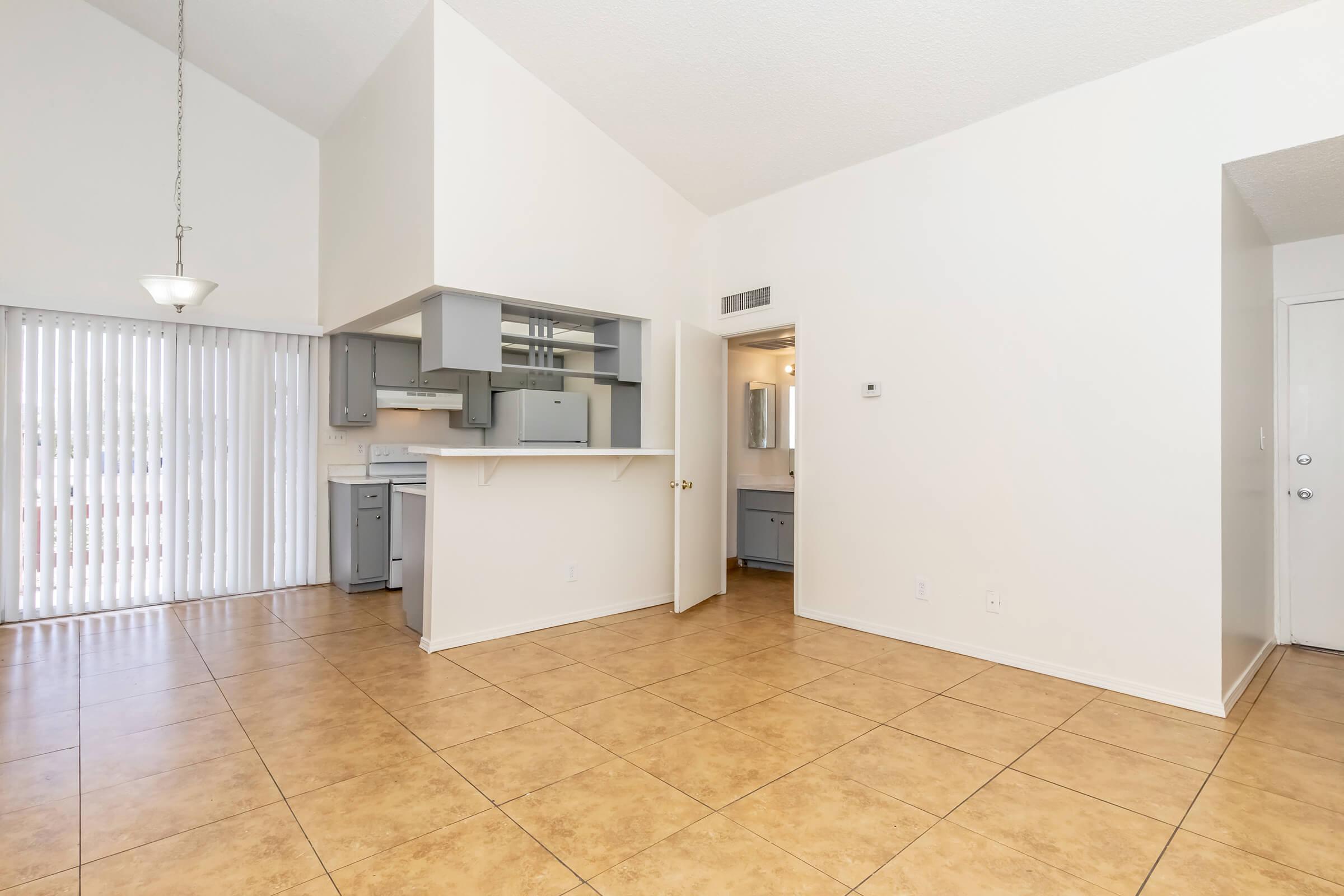 Bright, open living space with high ceilings, featuring beige tiled flooring. A kitchen area with light gray cabinets is visible in the background. To the right, a door leads to a bathroom, while a sliding door opens to a patio. Natural light floods the room, creating an inviting atmosphere.