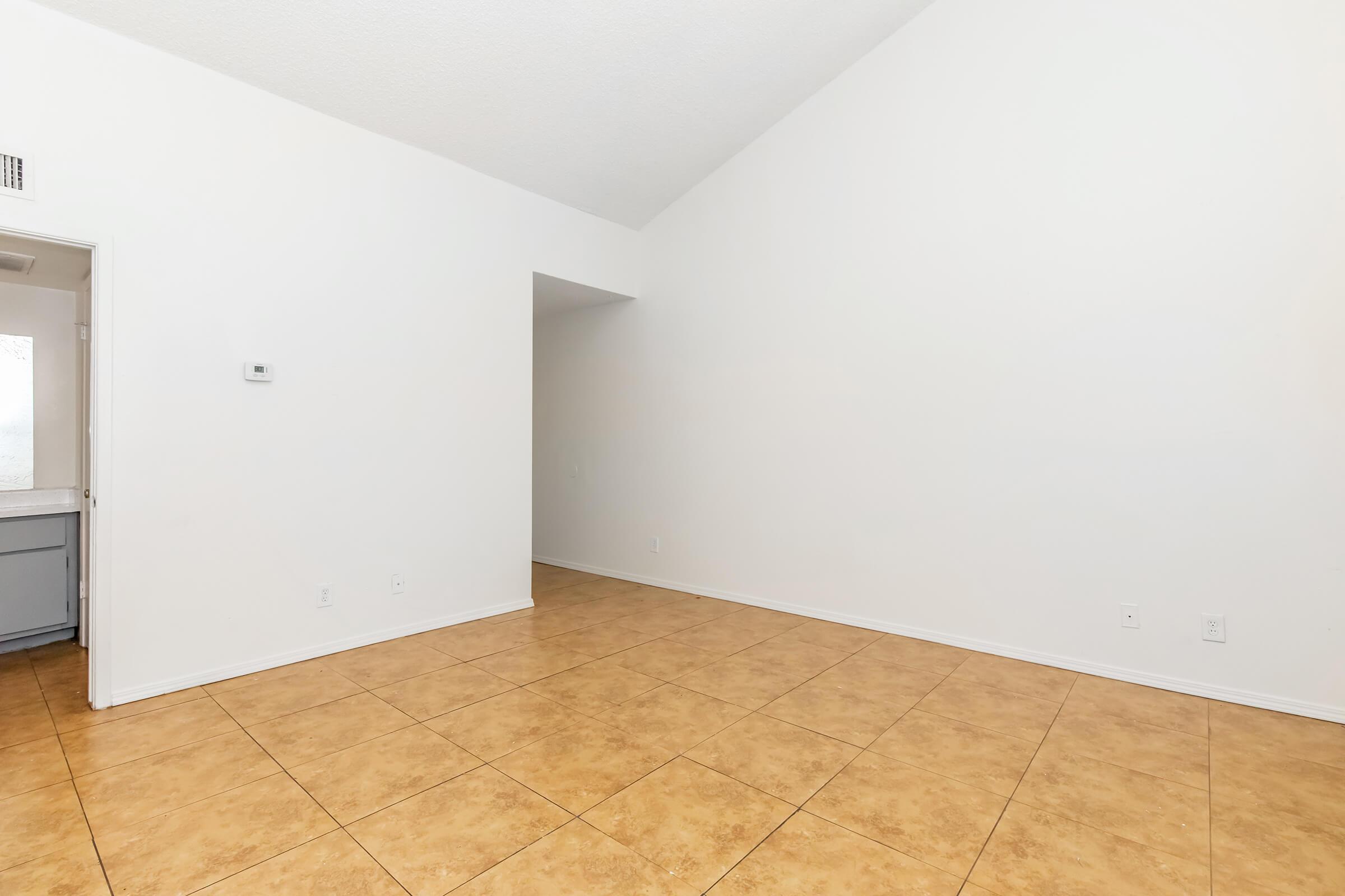 Spacious, sunlit room featuring light-colored walls and a high, vaulted ceiling. The floor is covered with light brown tiles, and there's an entryway leading into a kitchen area. The overall atmosphere is open and airy, making it suitable for various uses such as a living space or dining area.