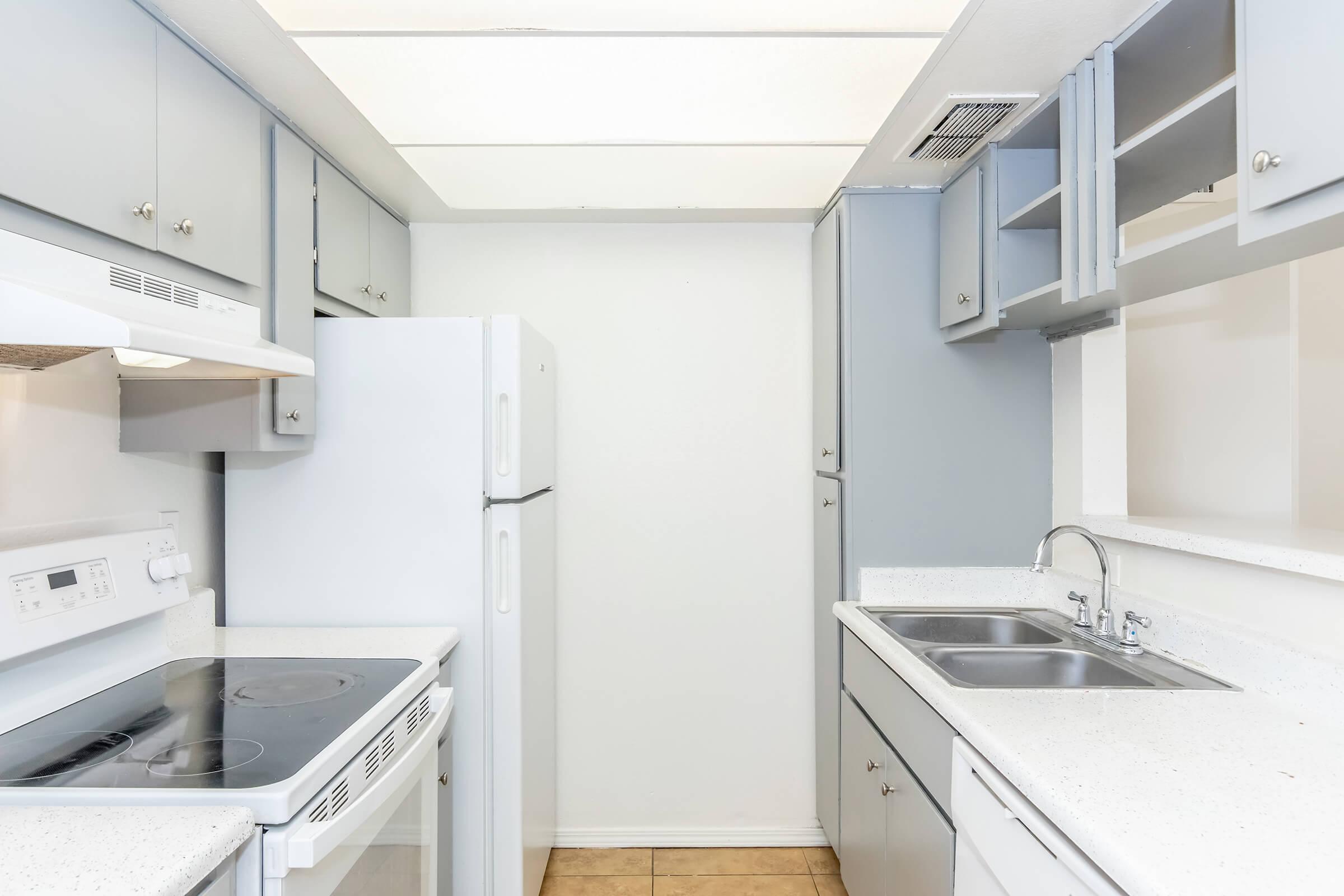 A modern kitchen featuring light gray cabinets, a white refrigerator, an electric stove with oven, a double sink, and a countertop with a speckled pattern. The space is well-lit with overhead lighting and has a clean, minimalist design.