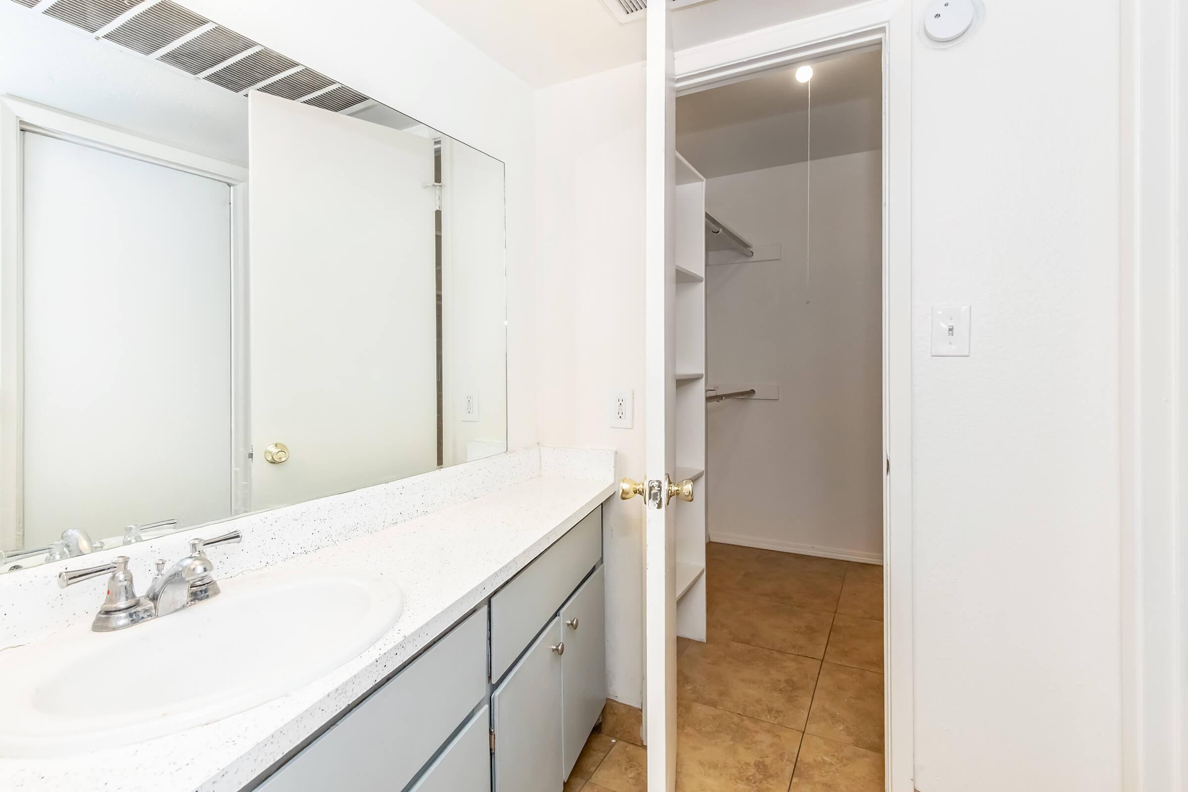 A bathroom featuring a countertop with a sink, a large mirror, and several cabinets. A doorway leads to a closet space with shelves. The walls are painted white, and the floor is tiled in a light color. Natural light is present from the ceiling fixture.