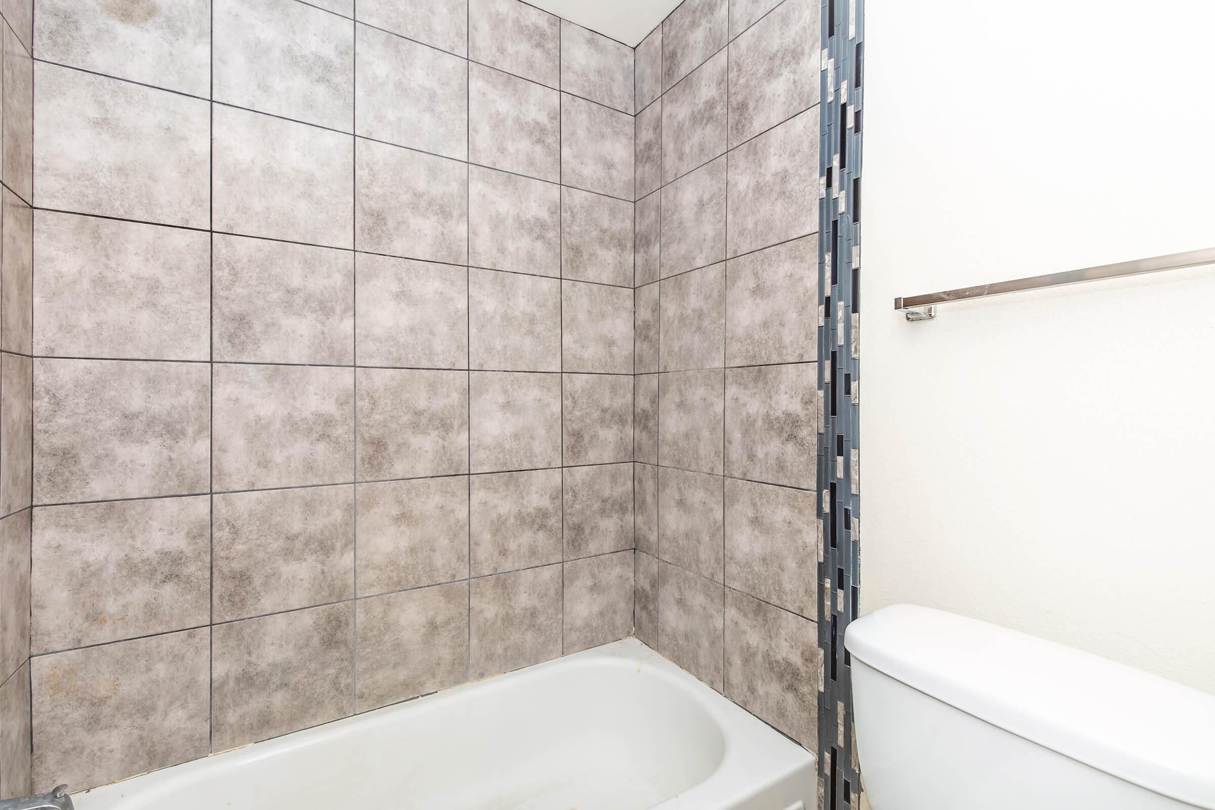 A bathroom with gray tiled walls, featuring a white bathtub and a toilet. The bathtub is empty, and the walls are covered in a light gray tile pattern. A chrome towel bar is mounted on the wall, and part of a glass shower enclosure is visible. The overall atmosphere is clean and simple.