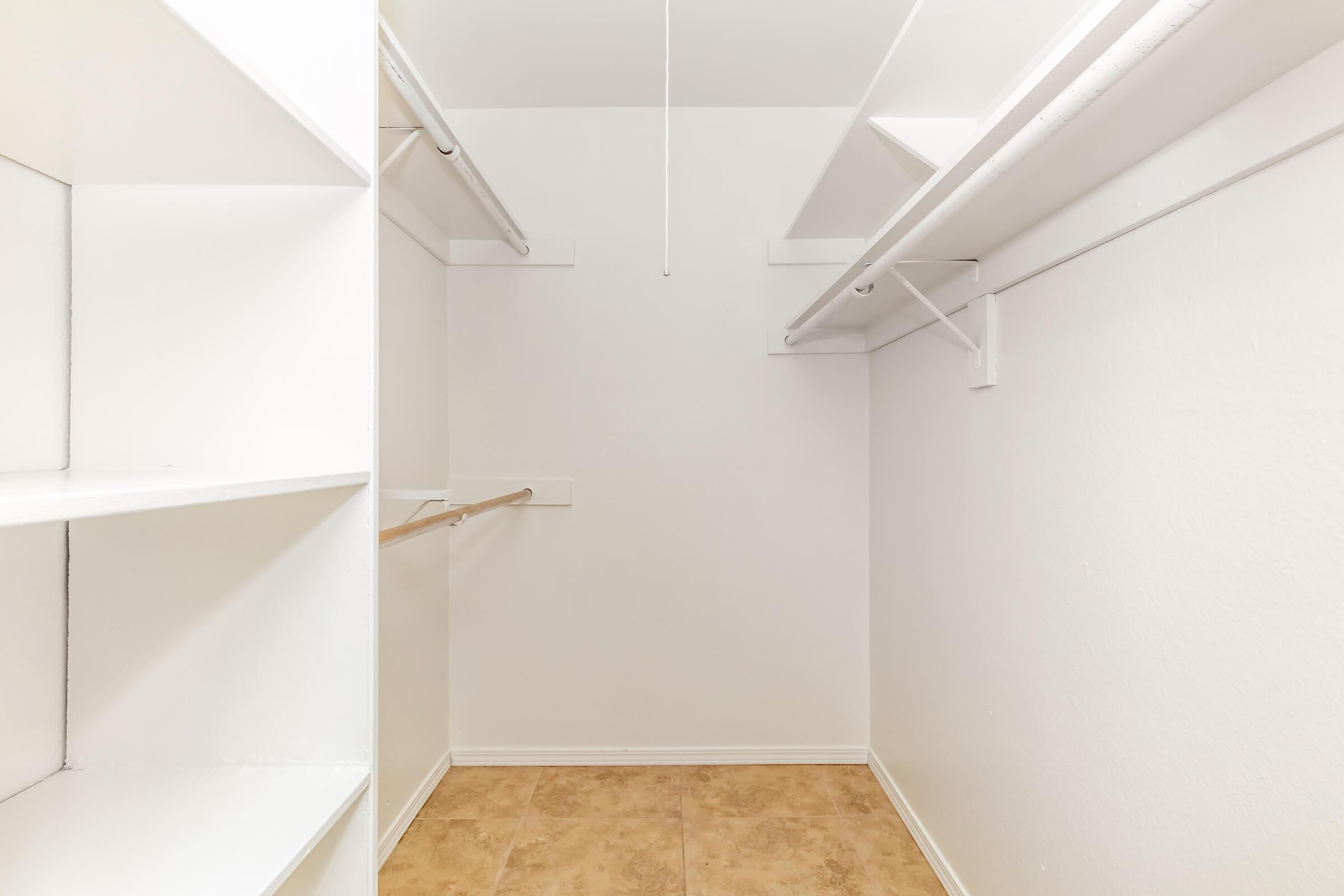 Empty closet space with white walls, featuring built-in shelving on the left side, a hanging rod for clothes in the center, and a tiled floor. The room is well-lit and includes a ceiling light fixture.
