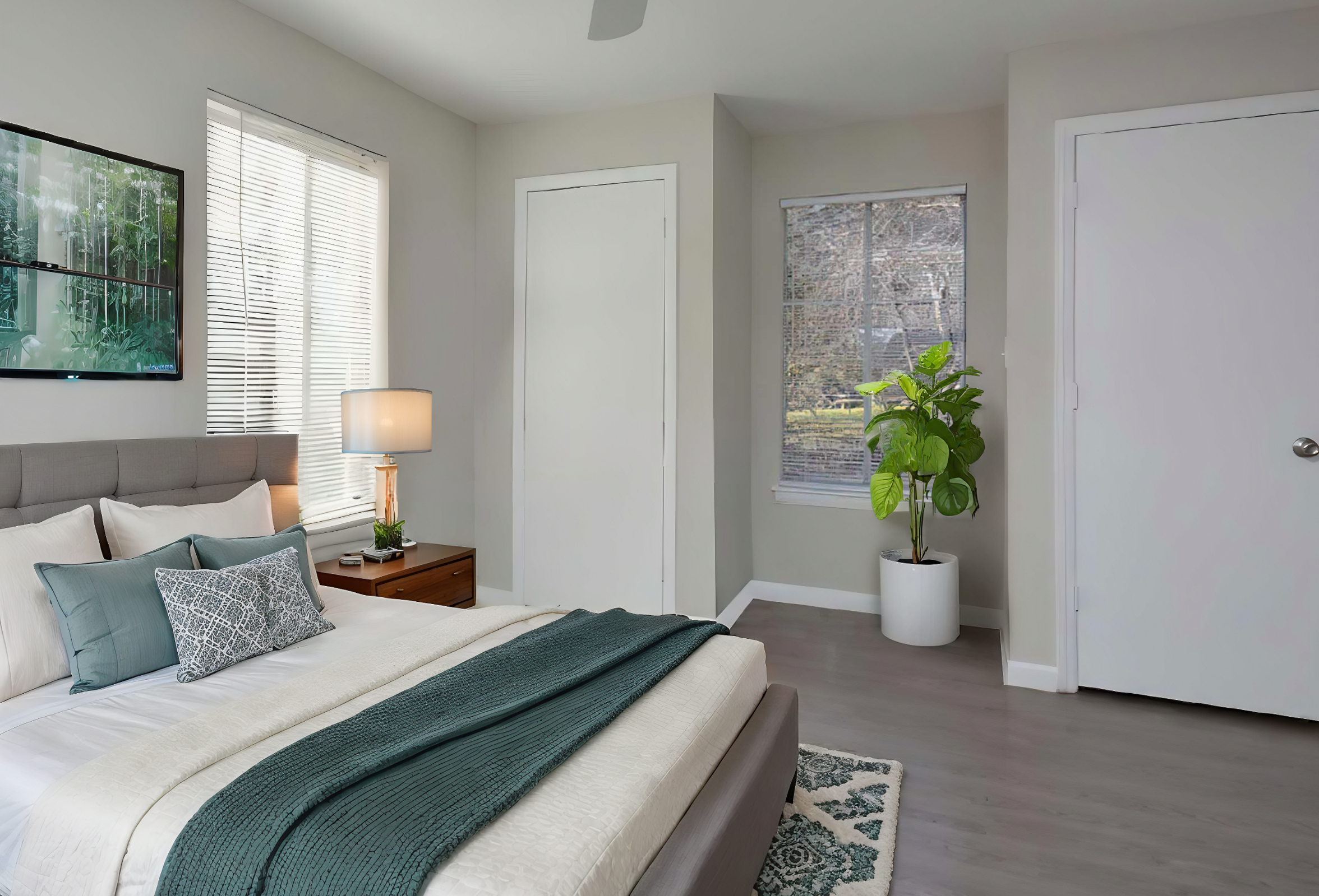 A cozy bedroom featuring a bed with a gray headboard and textured bedding, a bedside table with a lamp, and a framed nature artwork on the wall. Natural light enters through two windows, one with blinds and the other with a view of a plant. A potted plant adds a touch of greenery to the room.