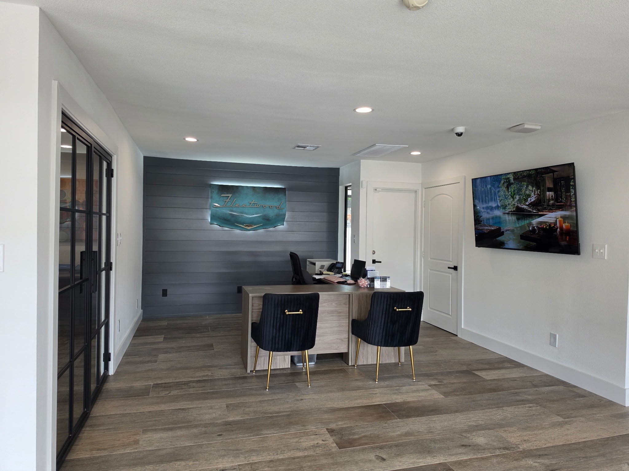 A modern reception area featuring a sleek desk with two black chairs, a decorative wall with a blue glowing sign, and a mounted flat-screen TV displaying a scenic landscape. The floor is made of wooden planks, and there's a glass door leading to another room. Bright, contemporary design elements create an inviting atmosphere.