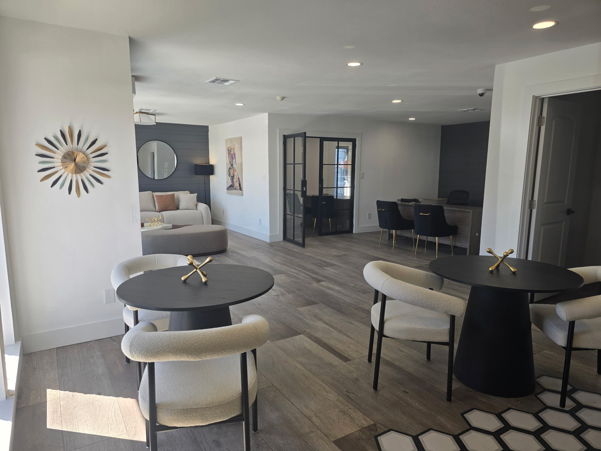 Modern interior of a spacious room featuring two round black tables with white upholstered chairs, a cozy seating area with a sofa, and a decorative wall clock. A glass partition leads to another section of the space, with stylish lighting and neutral tones creating an inviting atmosphere.