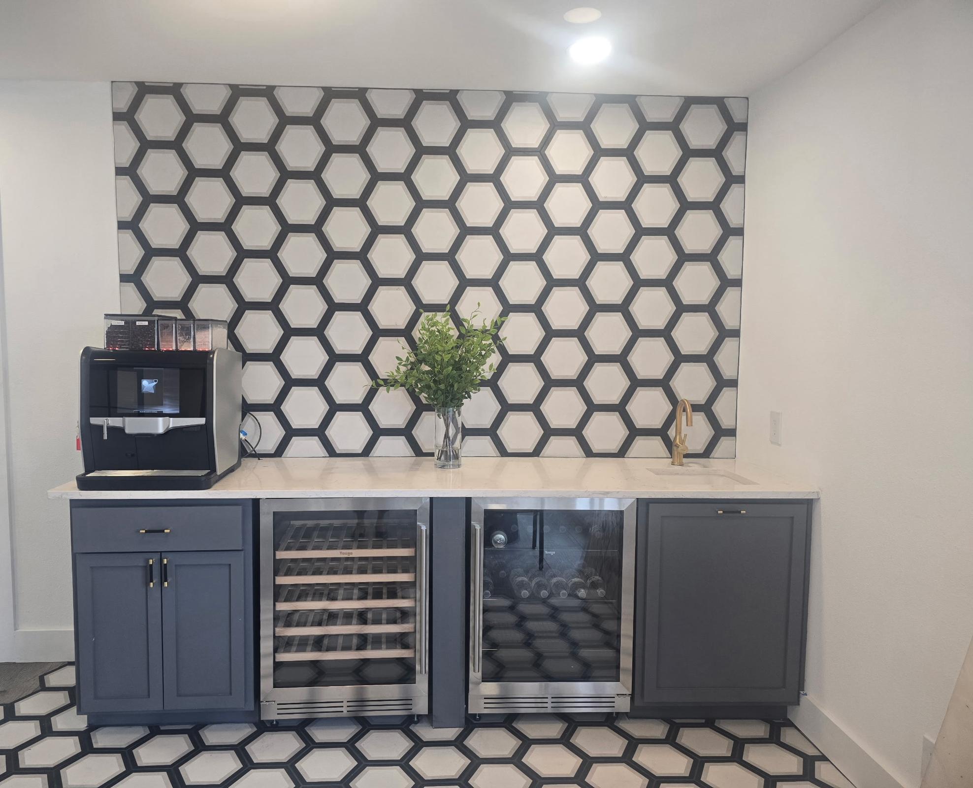 Modern kitchen area featuring a stylish hexagonal patterned wall, a sleek countertop, a coffee maker on the left, and two glass-front wine refrigerators below. A decorative vase with greenery adds a touch of nature to the contemporary design.