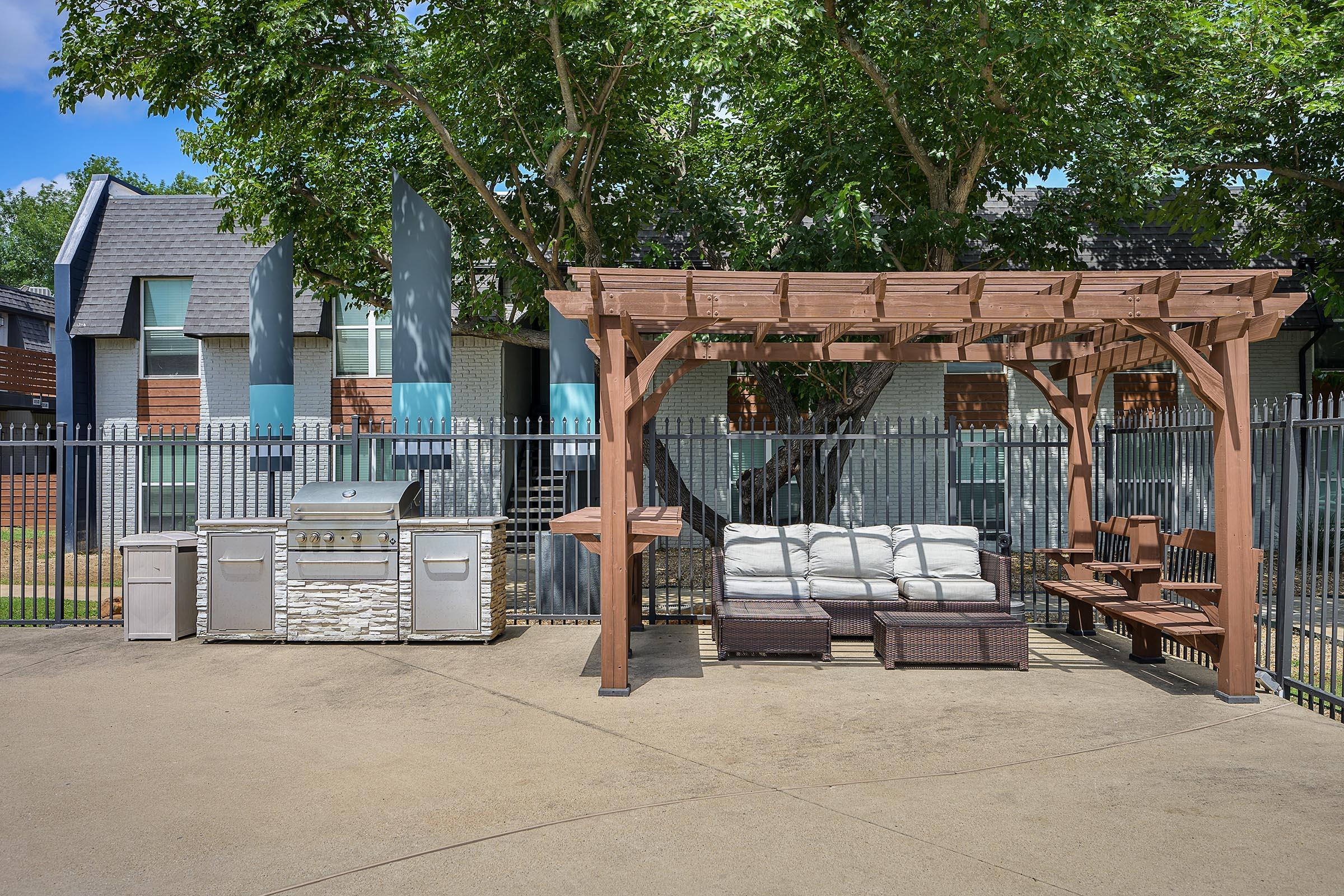 A backyard area featuring a wooden pergola with seating and a table, alongside a grill station with storage. The space is surrounded by a fence and trees, creating a shaded and inviting atmosphere for outdoor gatherings.