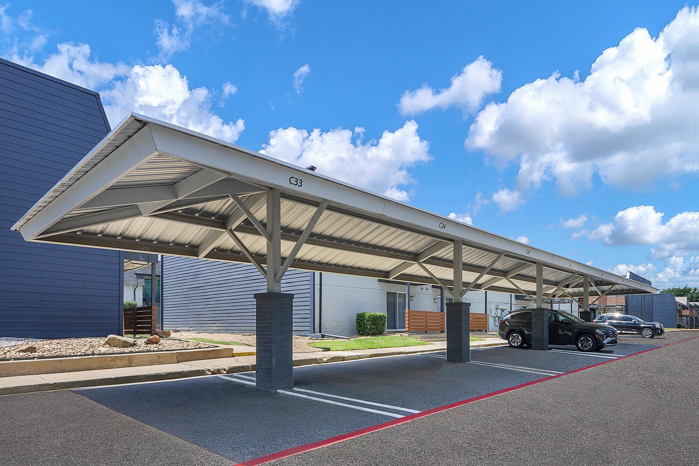 Carport structure with multiple parking spaces, featuring a modern design. It has a tall roof supported by steel beams, created for shade and protection from the elements. Clear blue sky with fluffy white clouds in the background, and a few parked cars visible beneath the carport in a parking lot.