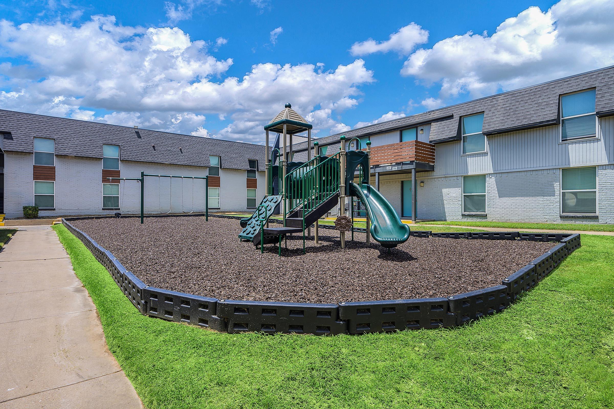 A playground with green play structures, including a slide and swings, situated on a grassy area surrounded by mulch. The backdrop features a multi-unit residential building under a blue sky with scattered clouds.