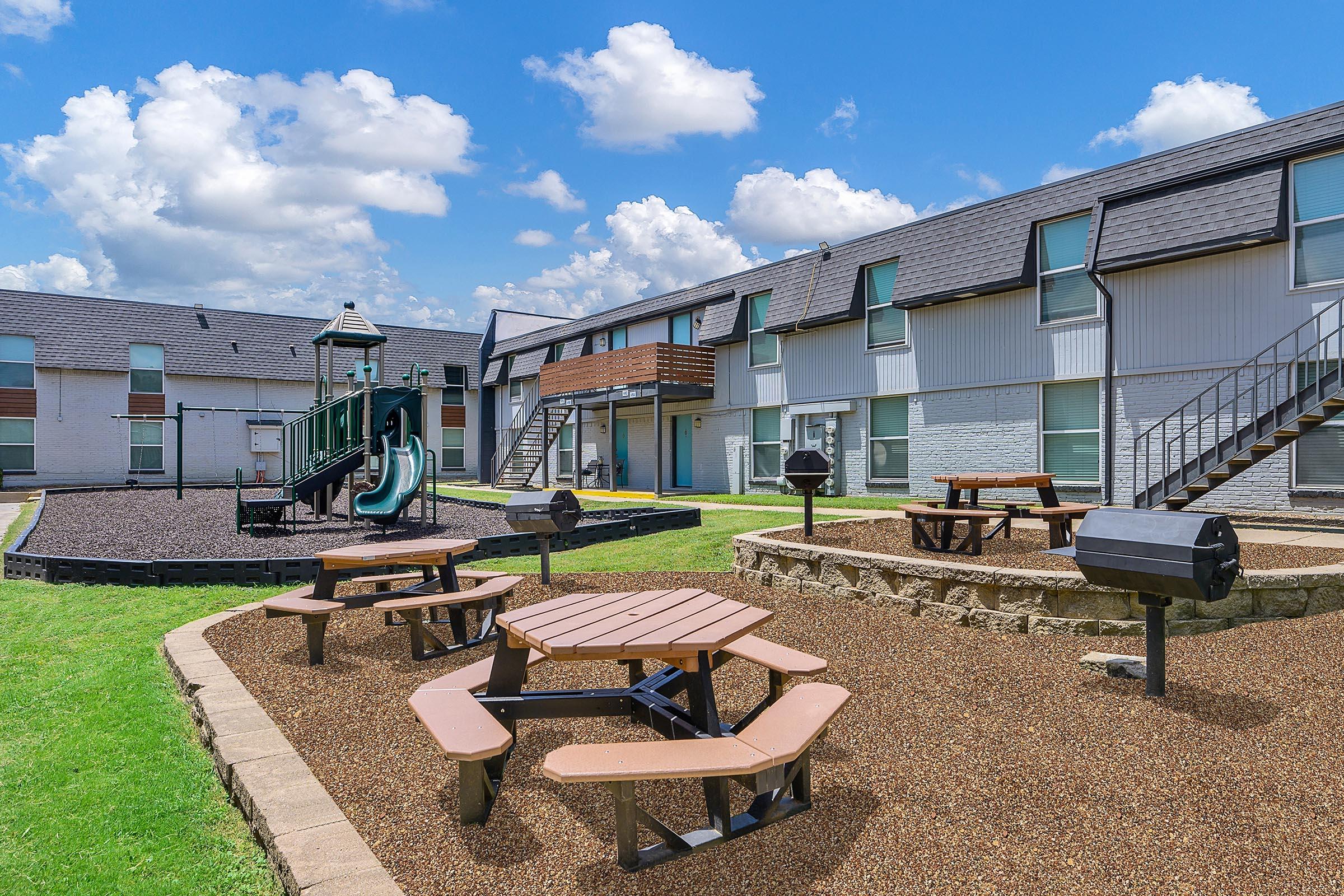 A sunny outdoor area featuring a playground with slides and climbing structures, surrounded by grassy space. There are several picnic tables and charcoal grills for barbecuing, with apartment buildings in the background and fluffy white clouds dotting the blue sky.
