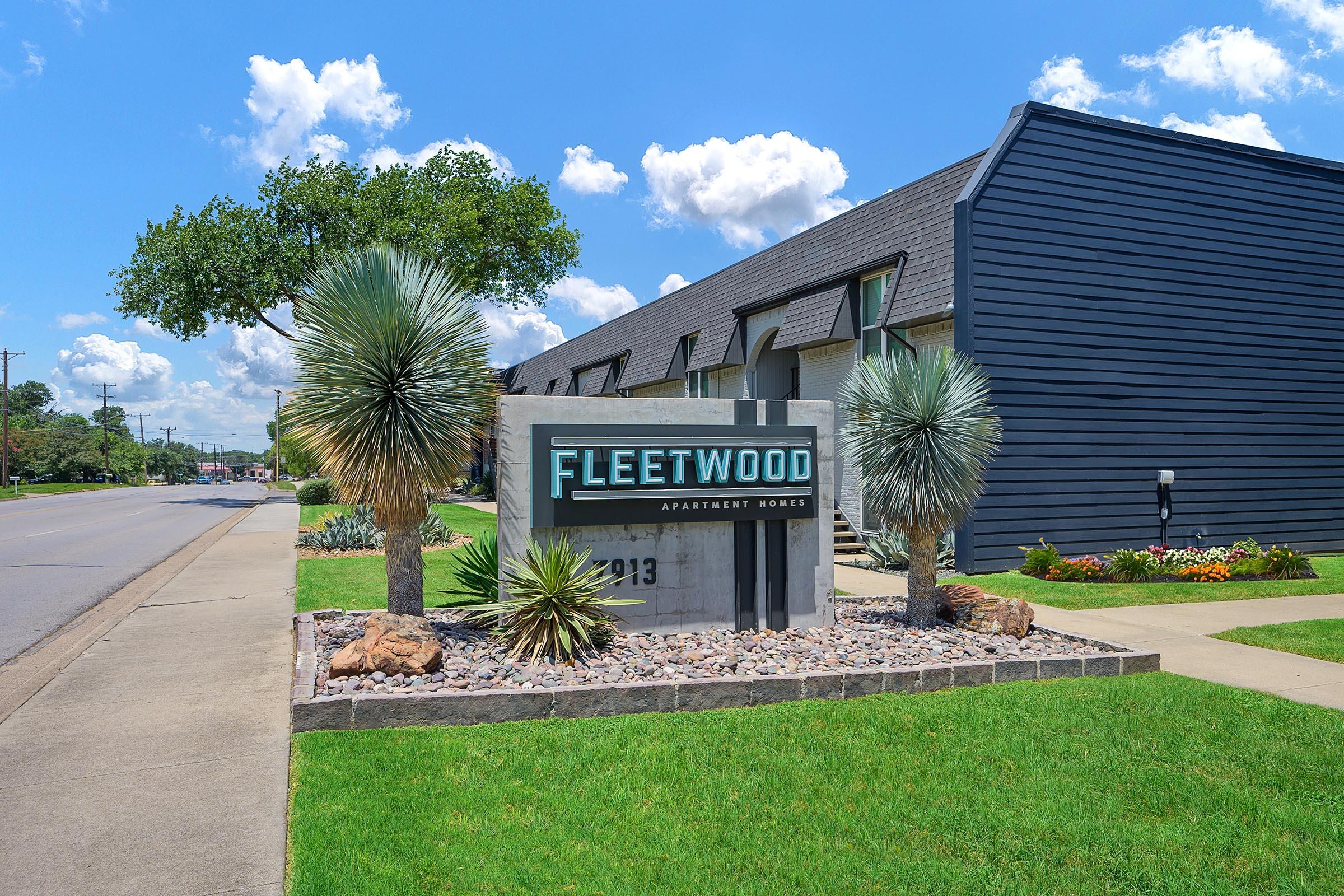 Exterior view of Fleetwood Apartment Homes, featuring a modern sign with the name prominently displayed. The landscape includes various types of rocks, shrubs, and palm trees, with a clear blue sky and fluffy clouds in the background. The building has a sleek, dark exterior with a sloped roof.